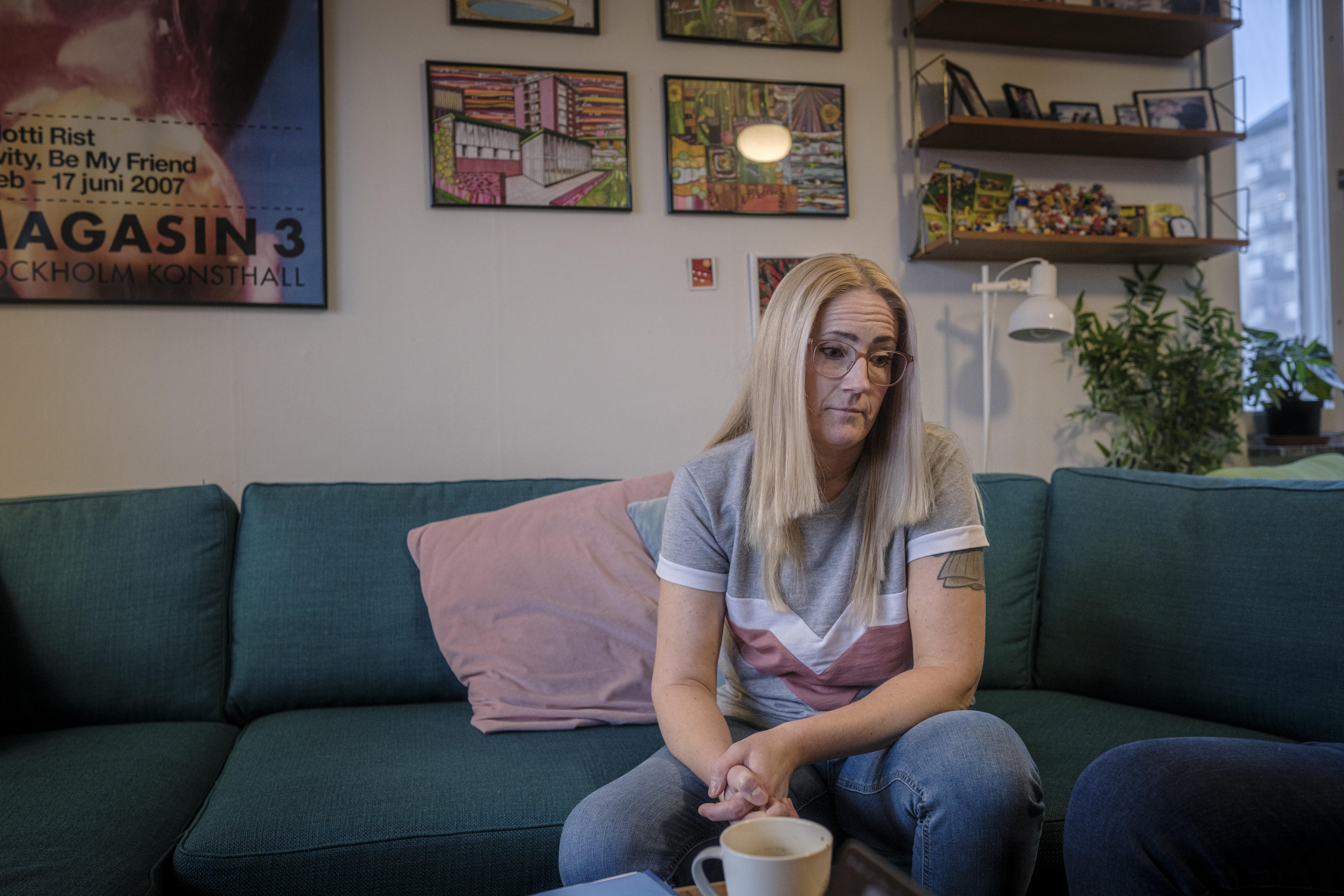 Karin Melin med långt blont hår och glasögon sitter i en soffa och dricker kaffe i ett rum med tavlor och växter.