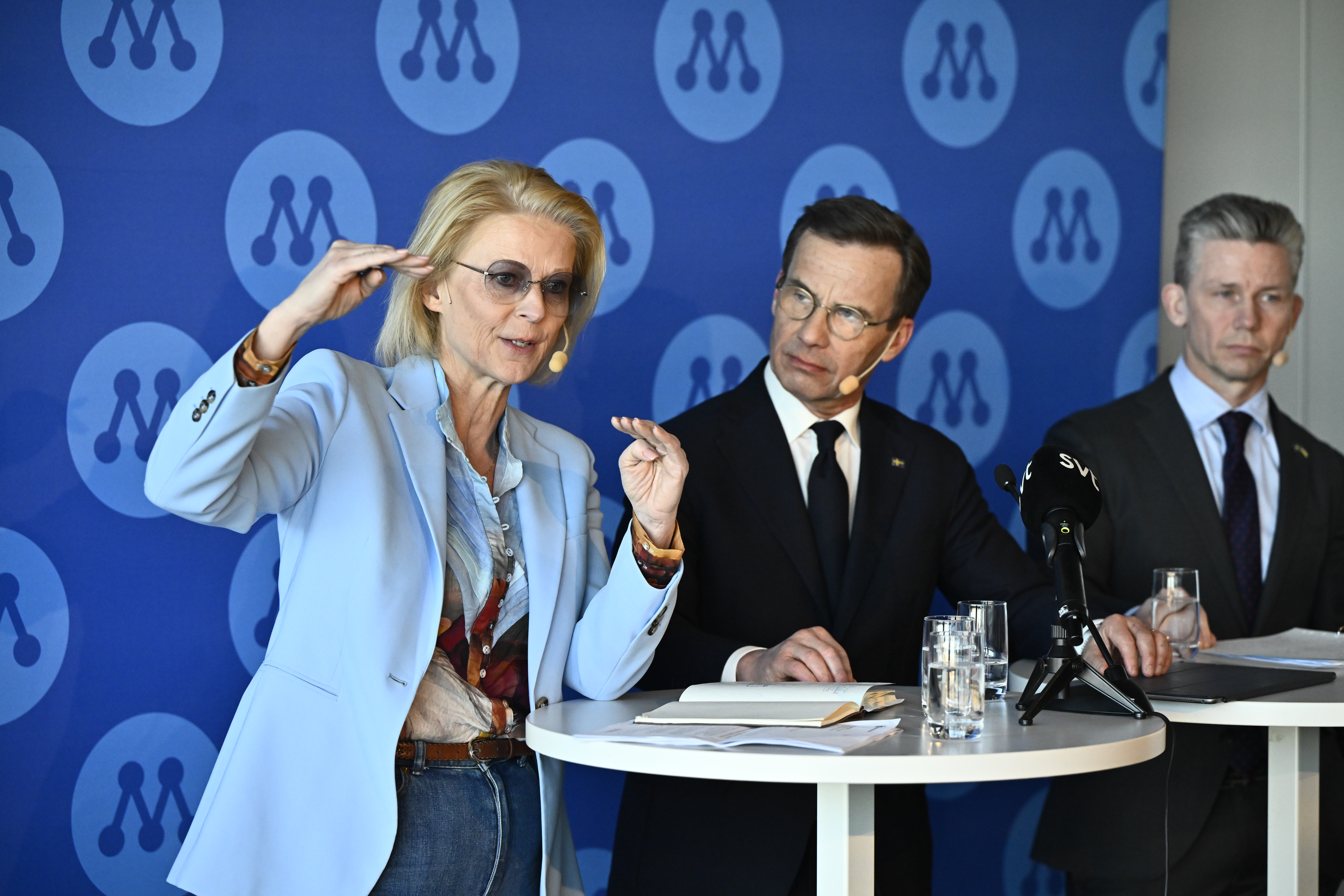 Två personer i formella kläder står vid ett runt bord med mikrofoner och vattenglas och talar framför en blå bakgrund med upprepade logotyper.