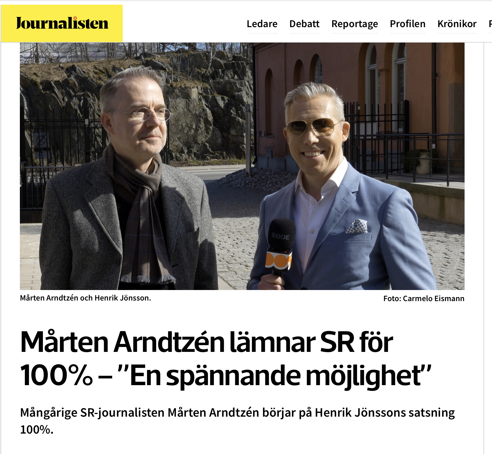 Två män står utomhus framför en byggnad med persikofärgad fasad. Den ena håller en mikrofon med orange logotyp.