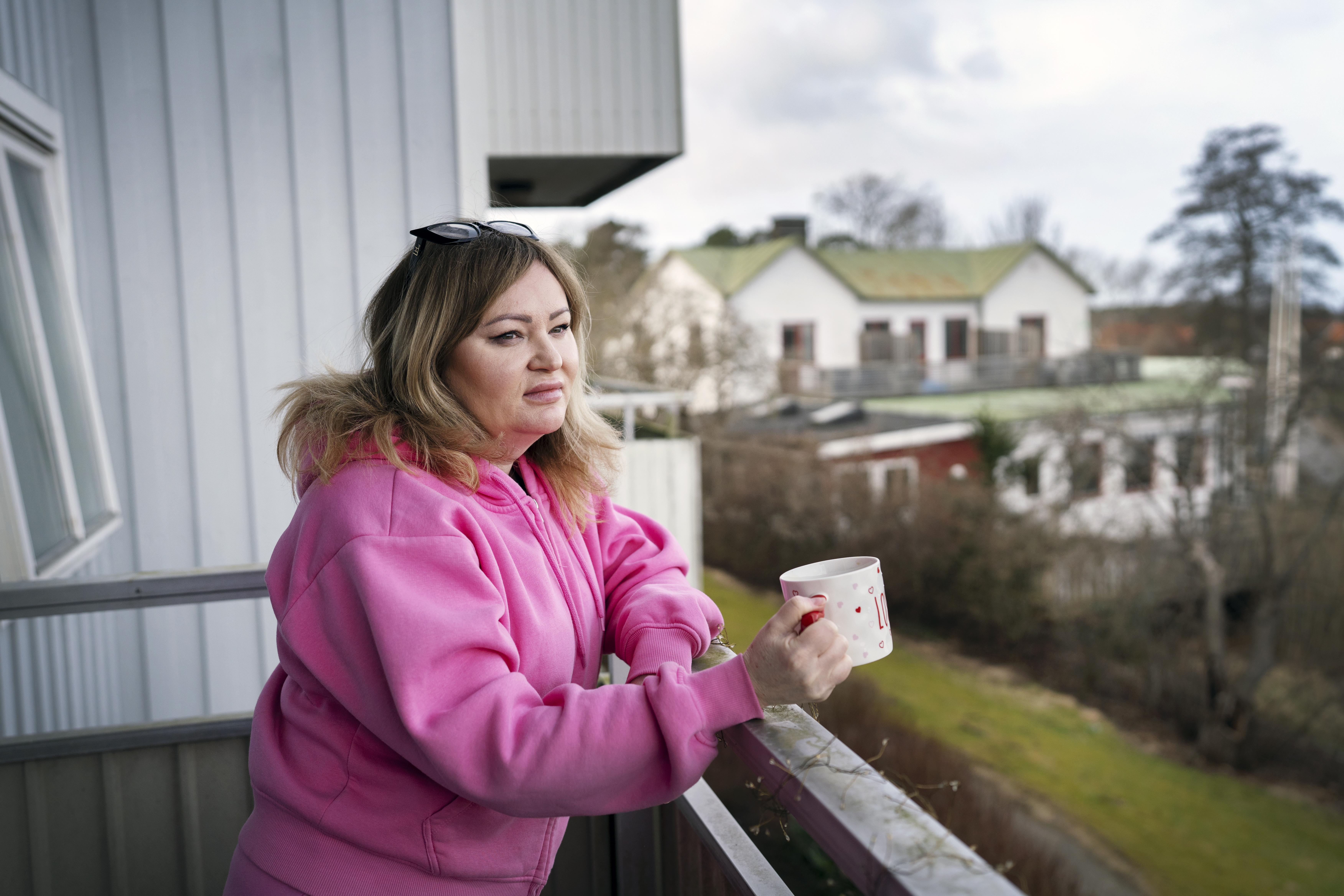 Olga i rosa huvtröja står på en balkong med en mugg och tittar ut över ett bostadsområde med hus och grönska.
