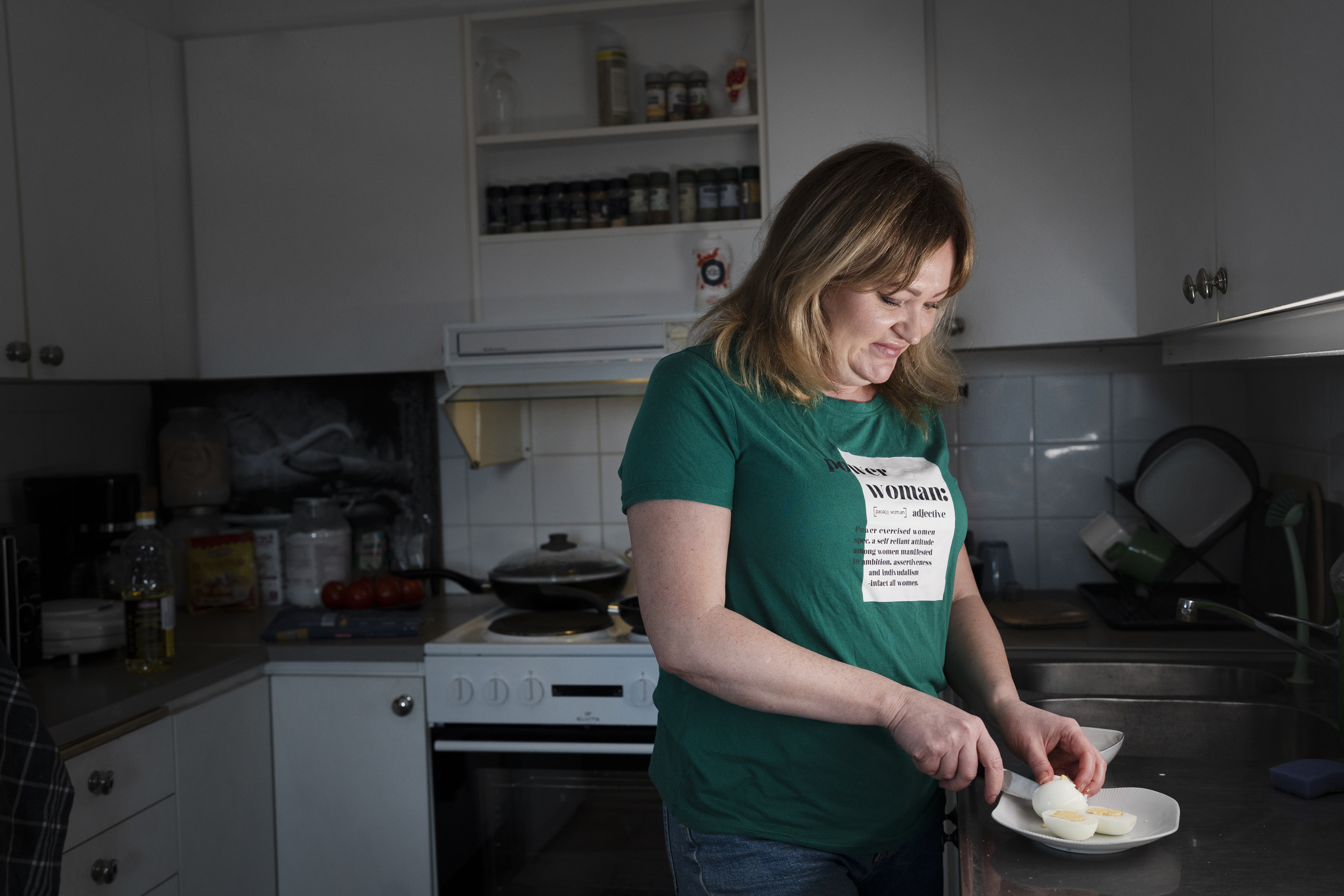 En kvinna står i köket och skivar ett kokt ägg på en tallrik. I bakgrunden syns hyllor med kryddor och köksmaskiner.