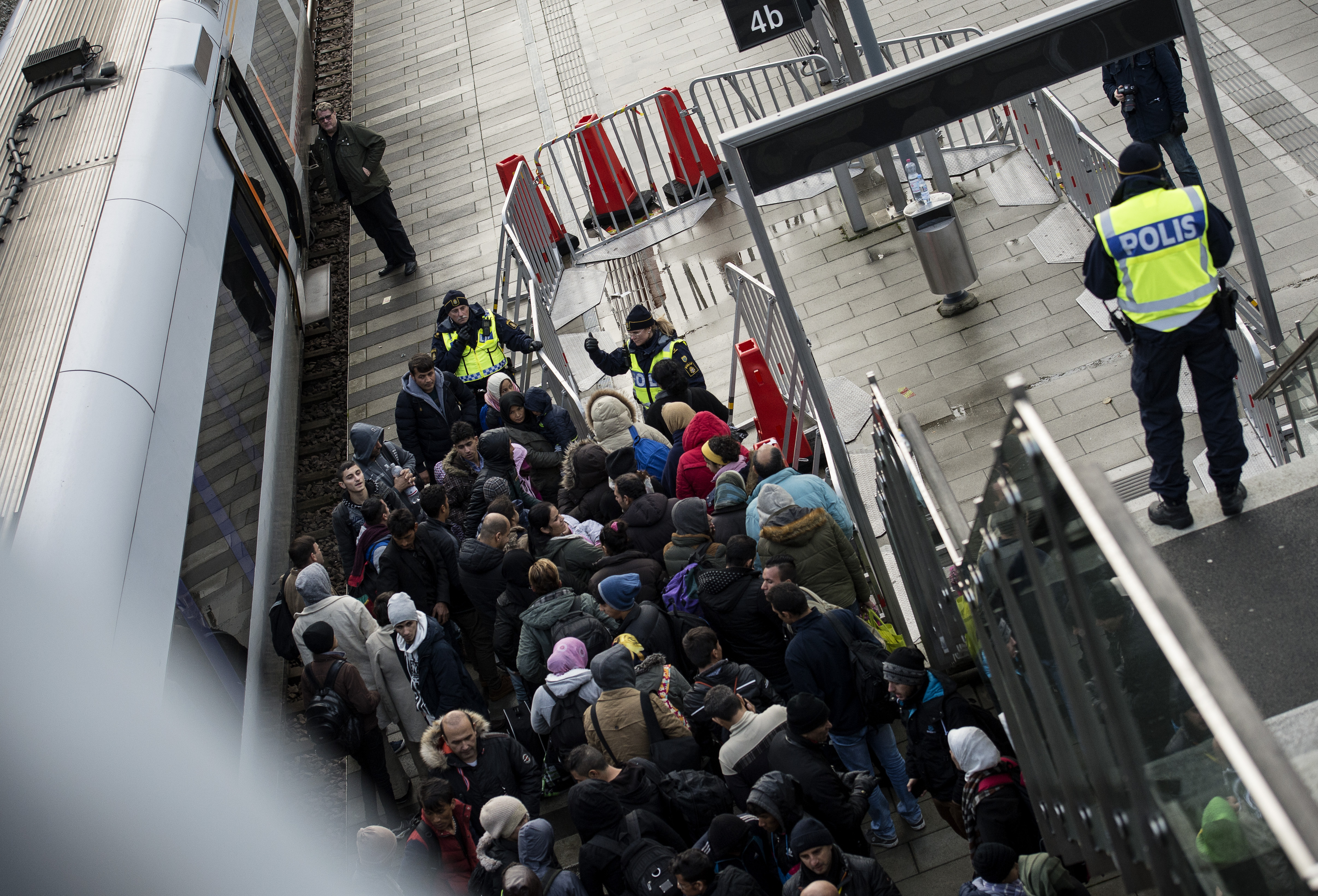 En grupp människor väntar på en tågperrong, övervakade av poliser i uniform bakom avspärrningar.