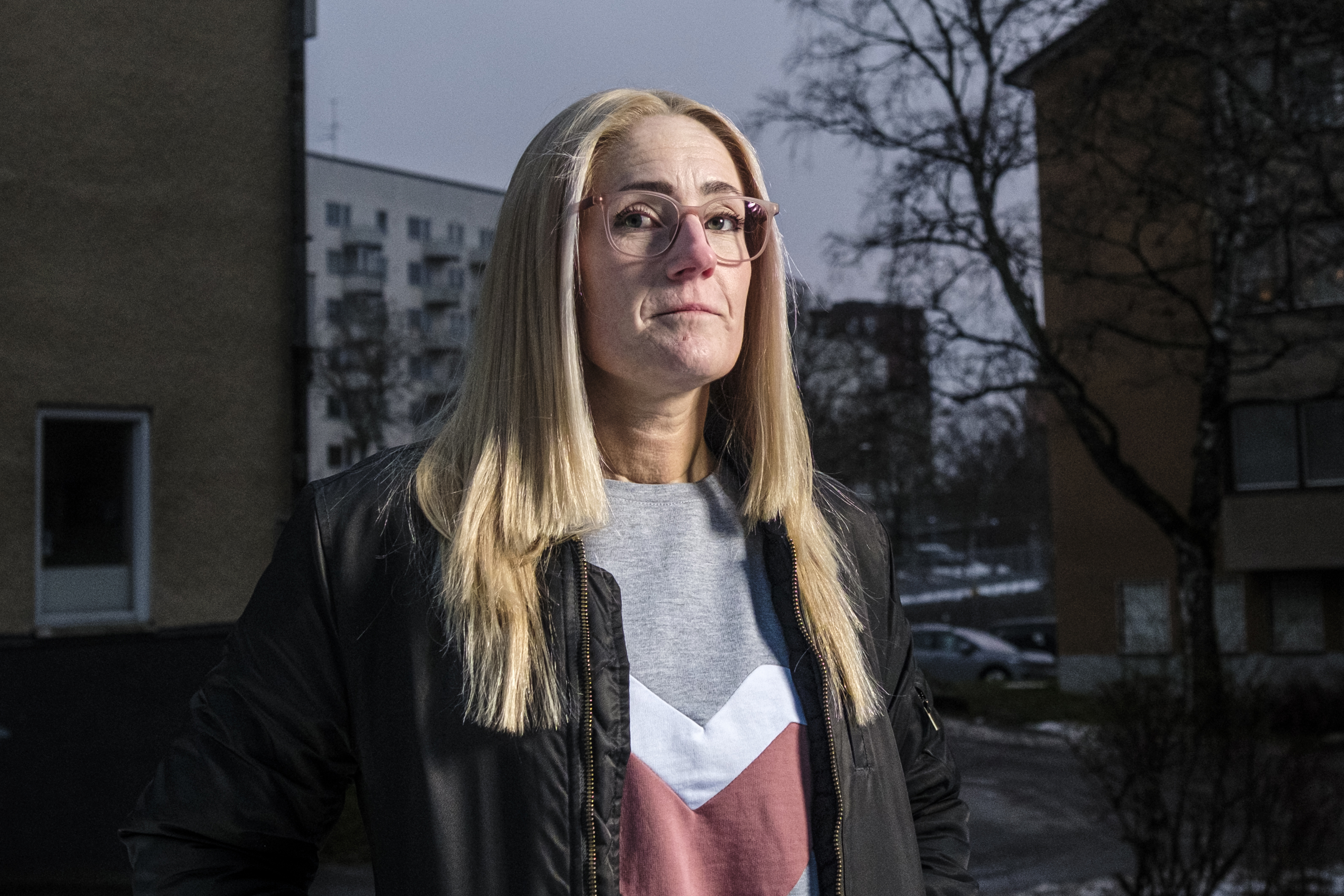Karin Melin, med långt blont hår och glasögon, står utomhus i ett bostadsområde iklädd kavaj och tröja. Du kan se hus och träd i bakgrunden.