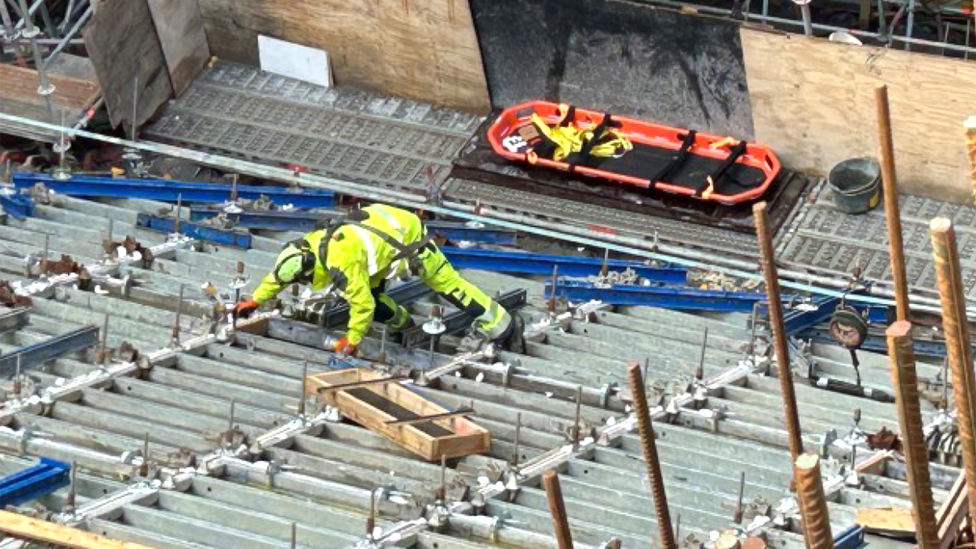 En byggarbetare i skyddsutrustning arbetar på en metallram vid en byggarbetsplats, med en bår synlig i närheten.