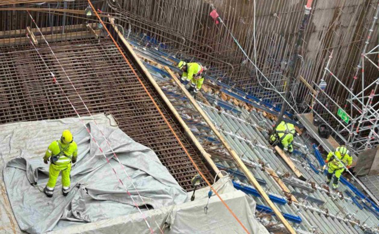 Fyra byggarbetare i varselkläder arbetar på en betongkonstruktion med stålarmering. Byggställning och utrustning syns.