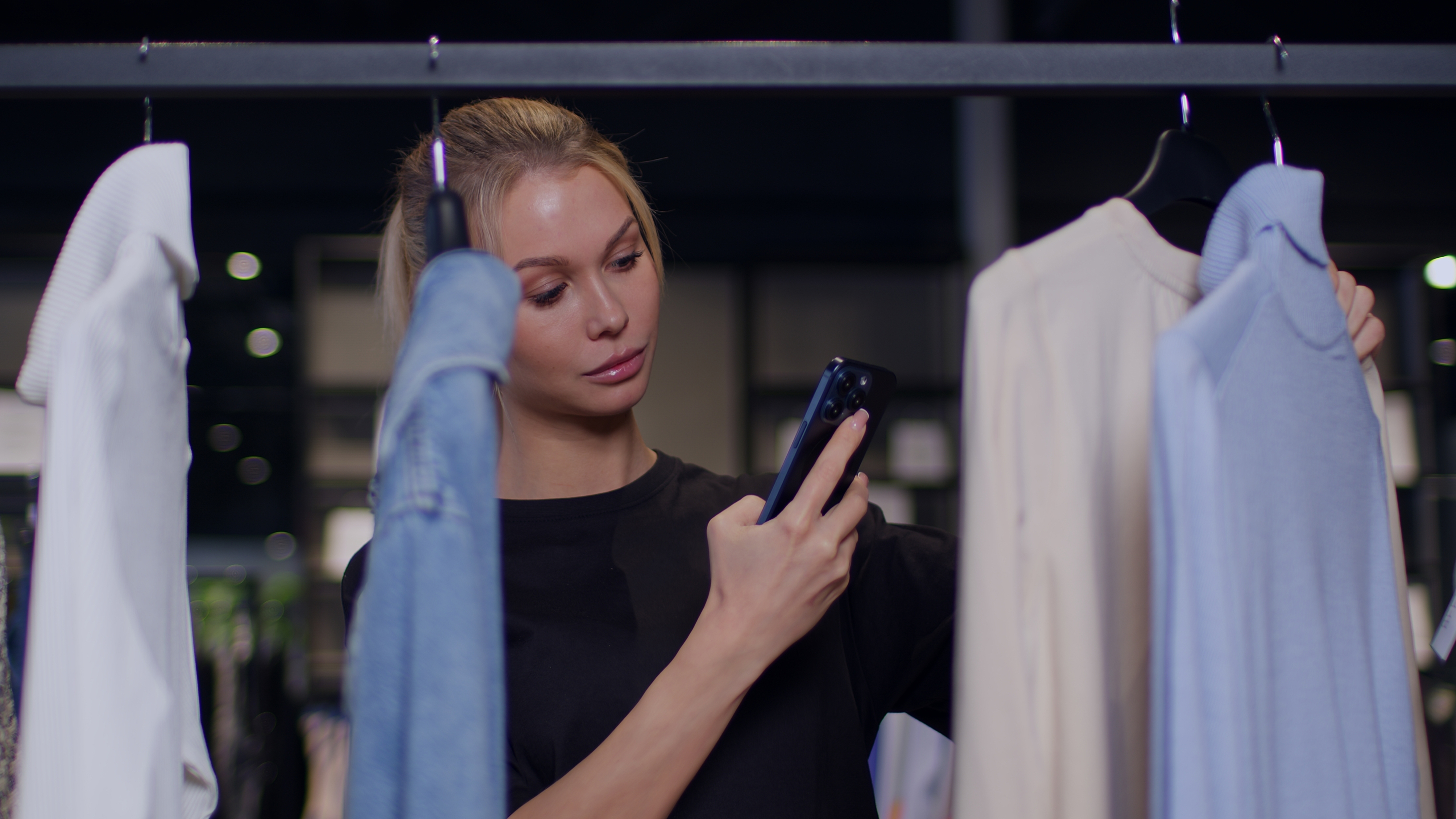 En kvinna håller en smartphone och tittar på kläder på galgar, mellan en blå skjorta och en beige tröja i en butik.