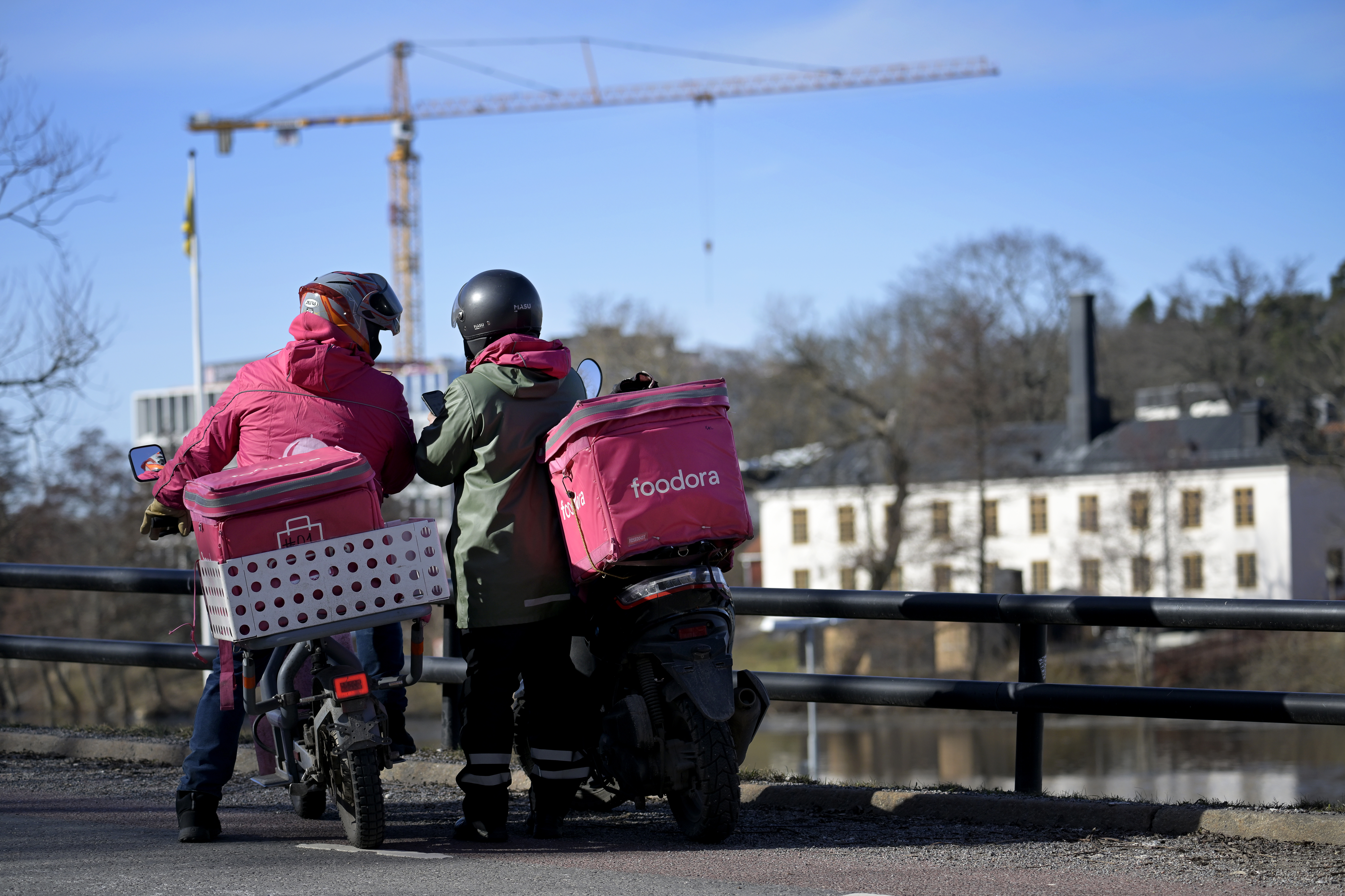 Två bud med Foodora-jackor och hjälmar står vid sina mopeder på en väg, med en byggkran och byggnader i bakgrunden.