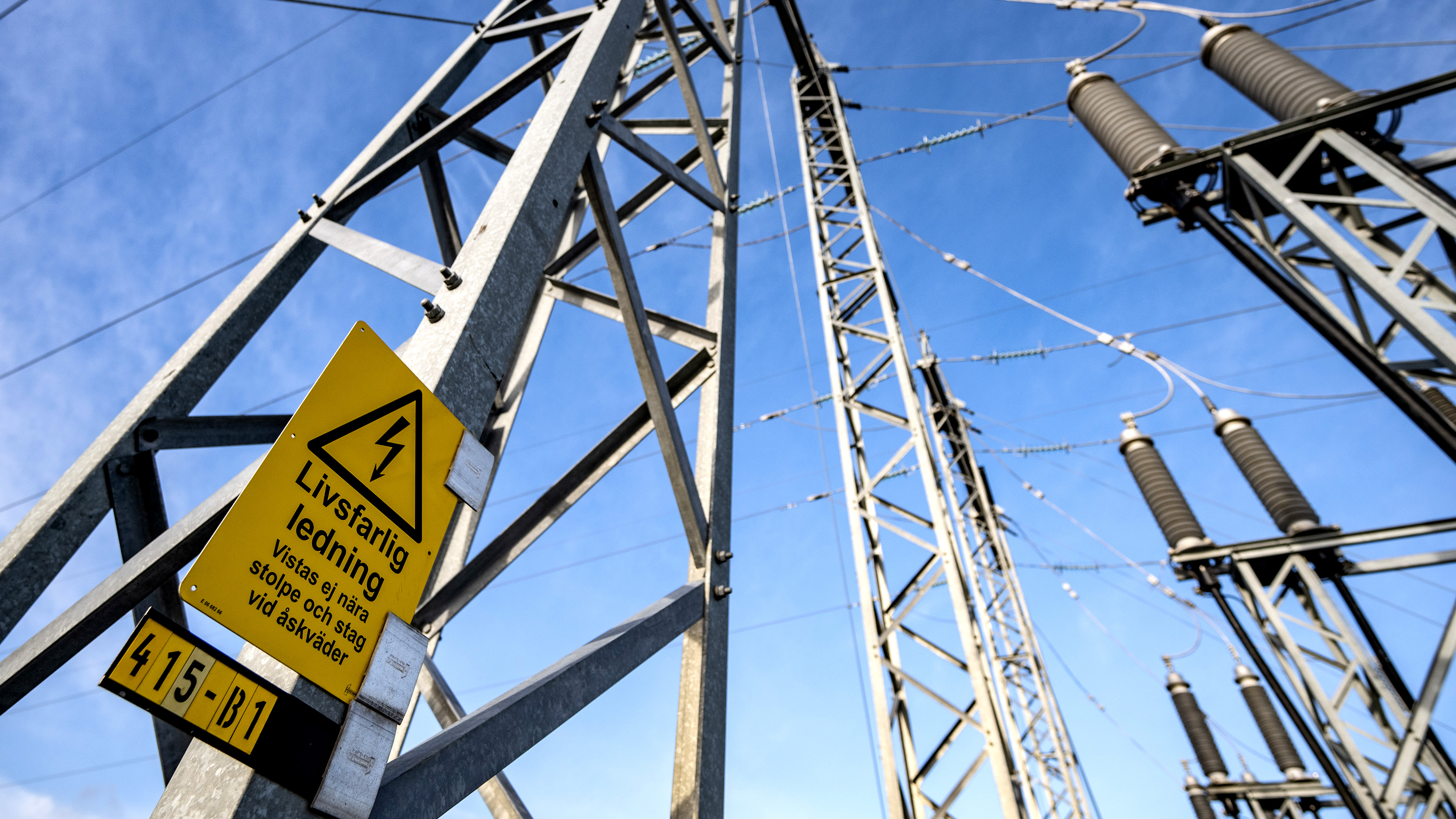Höga kraftledningsstolpar och ledningar under en himmel, med en gul varningsskylt på svenska om fara från elektriska ledningar.