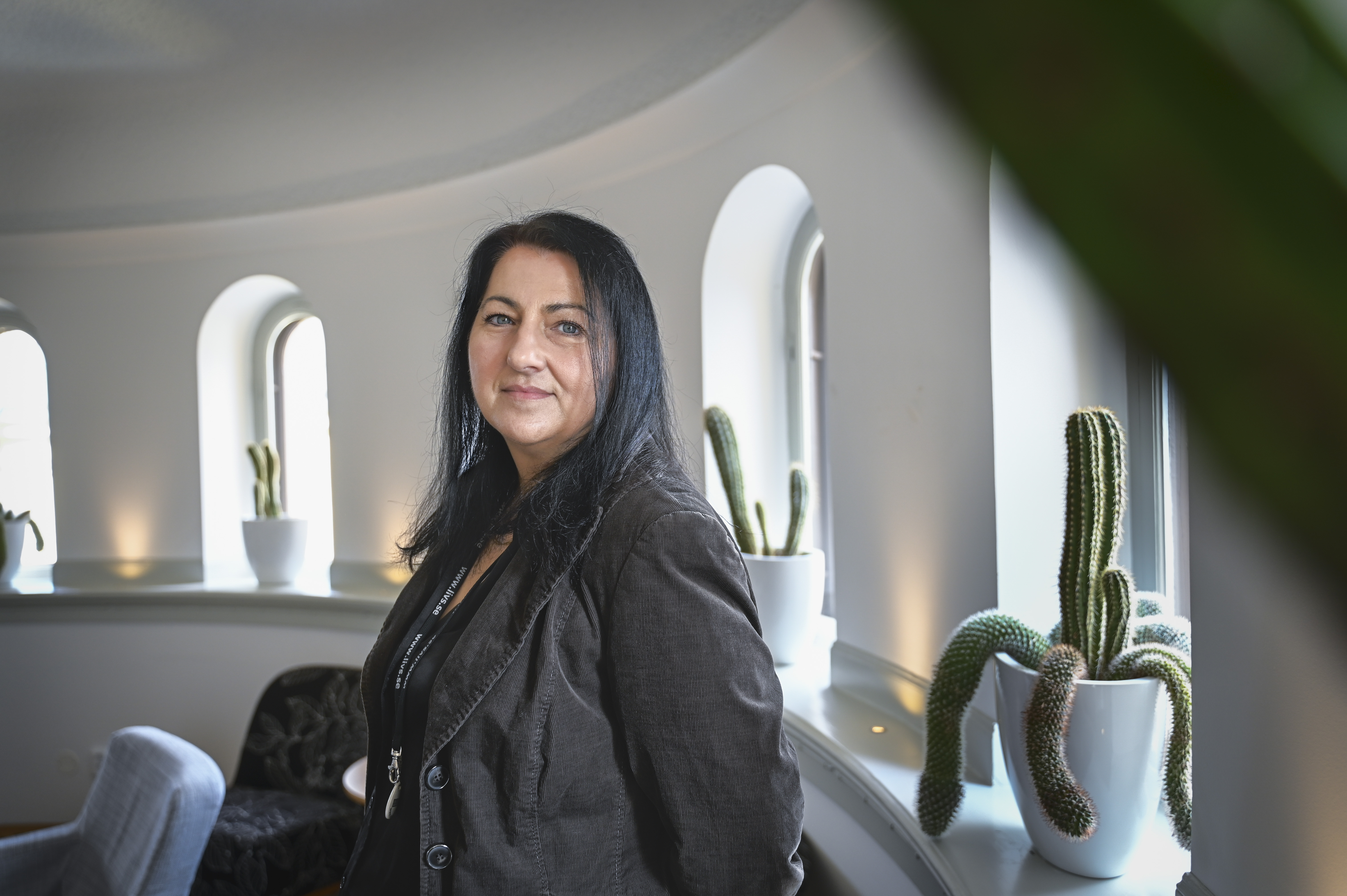 En kvinna med långt mörkt hår står inomhus nära välvda fönster och krukväxter, har en mörk jacka och ler svagt.