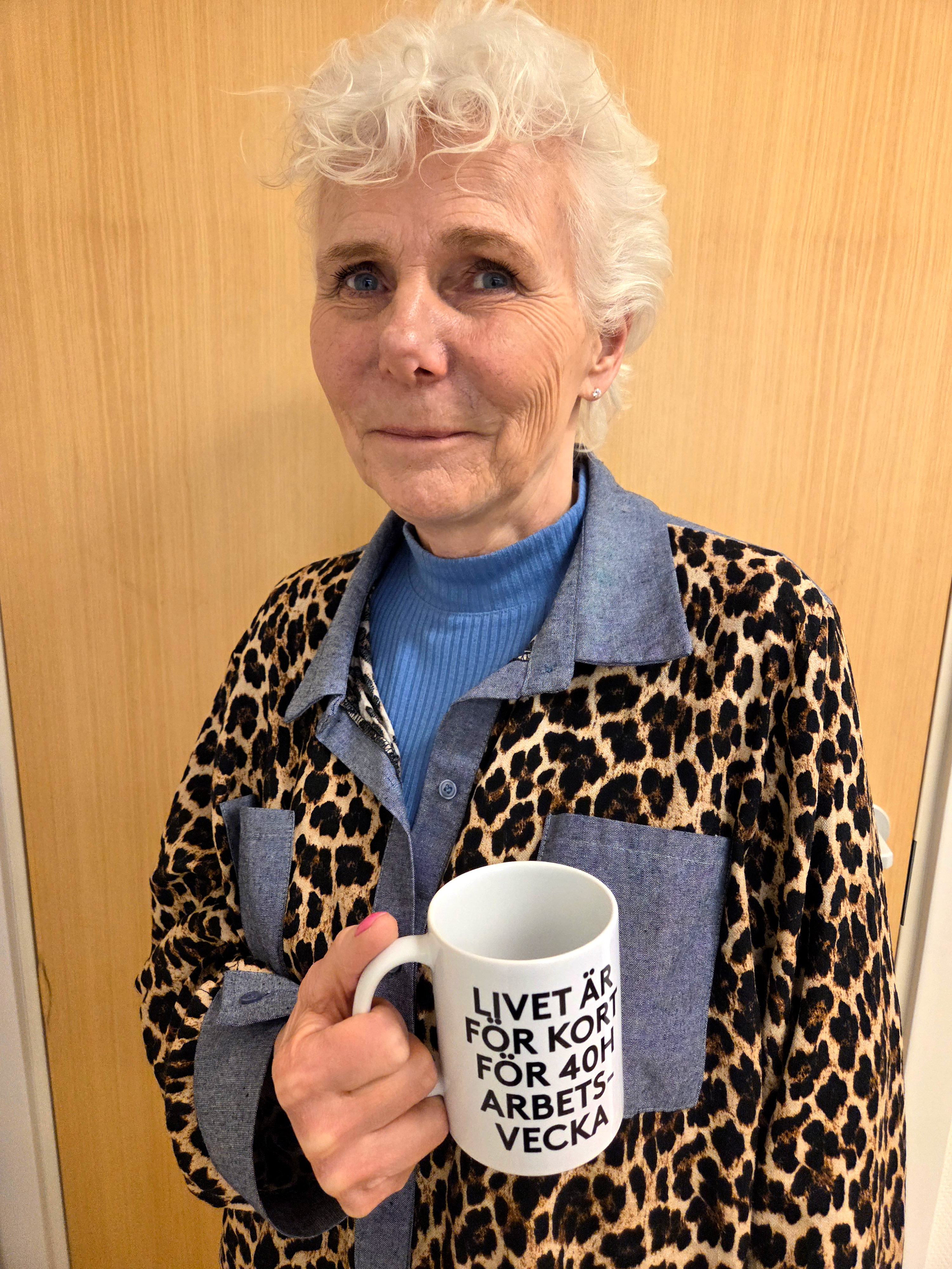 En äldre kvinna med kort vitt hår har på sig en leopardmönstrad jacka och håller en mugg med svensk text vid en trädörr.