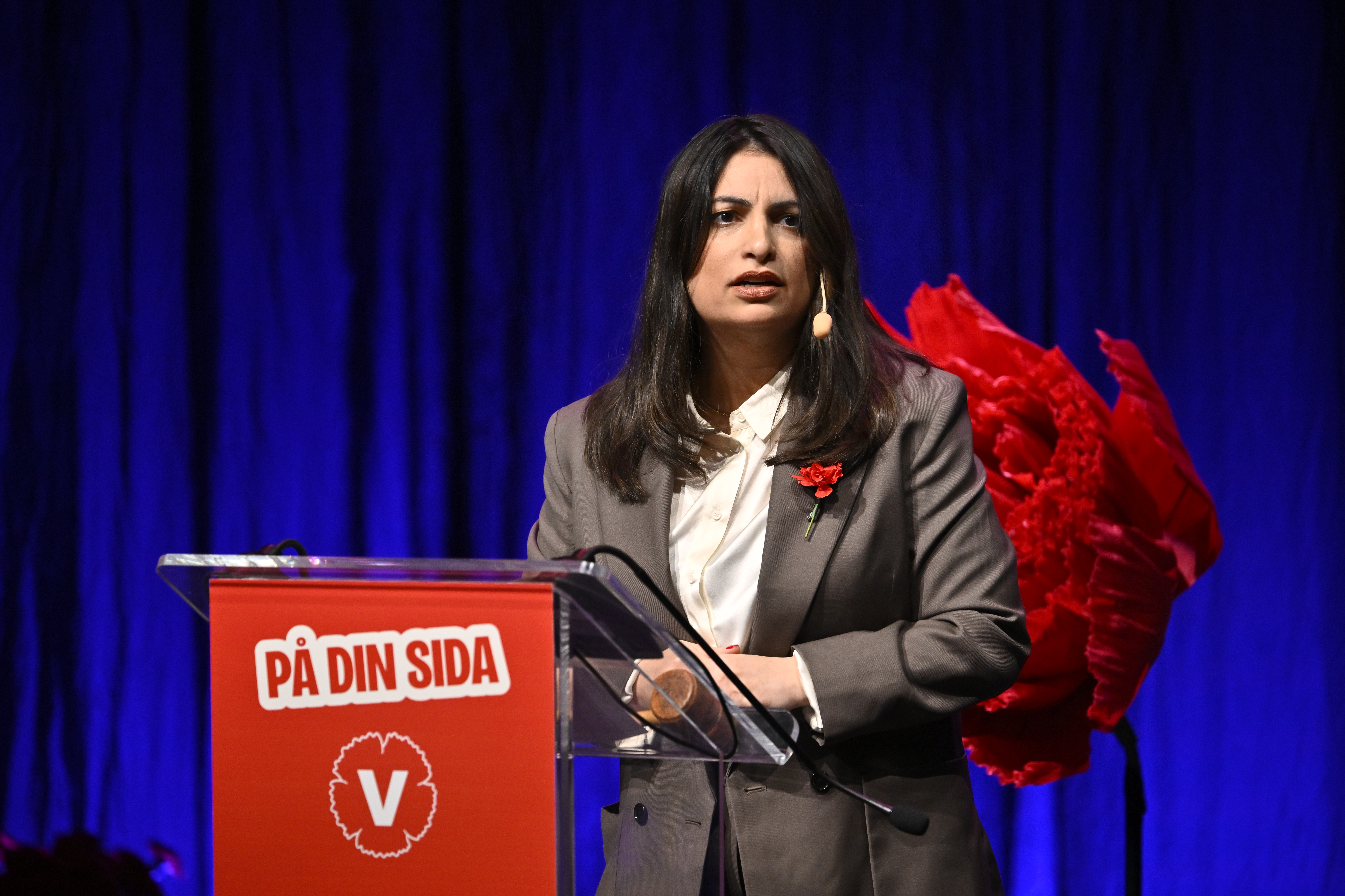 En kvinna i kostym står vid ett podium med texten "På din sida V", talar i headset och har en röd blomma på sin kavaj.