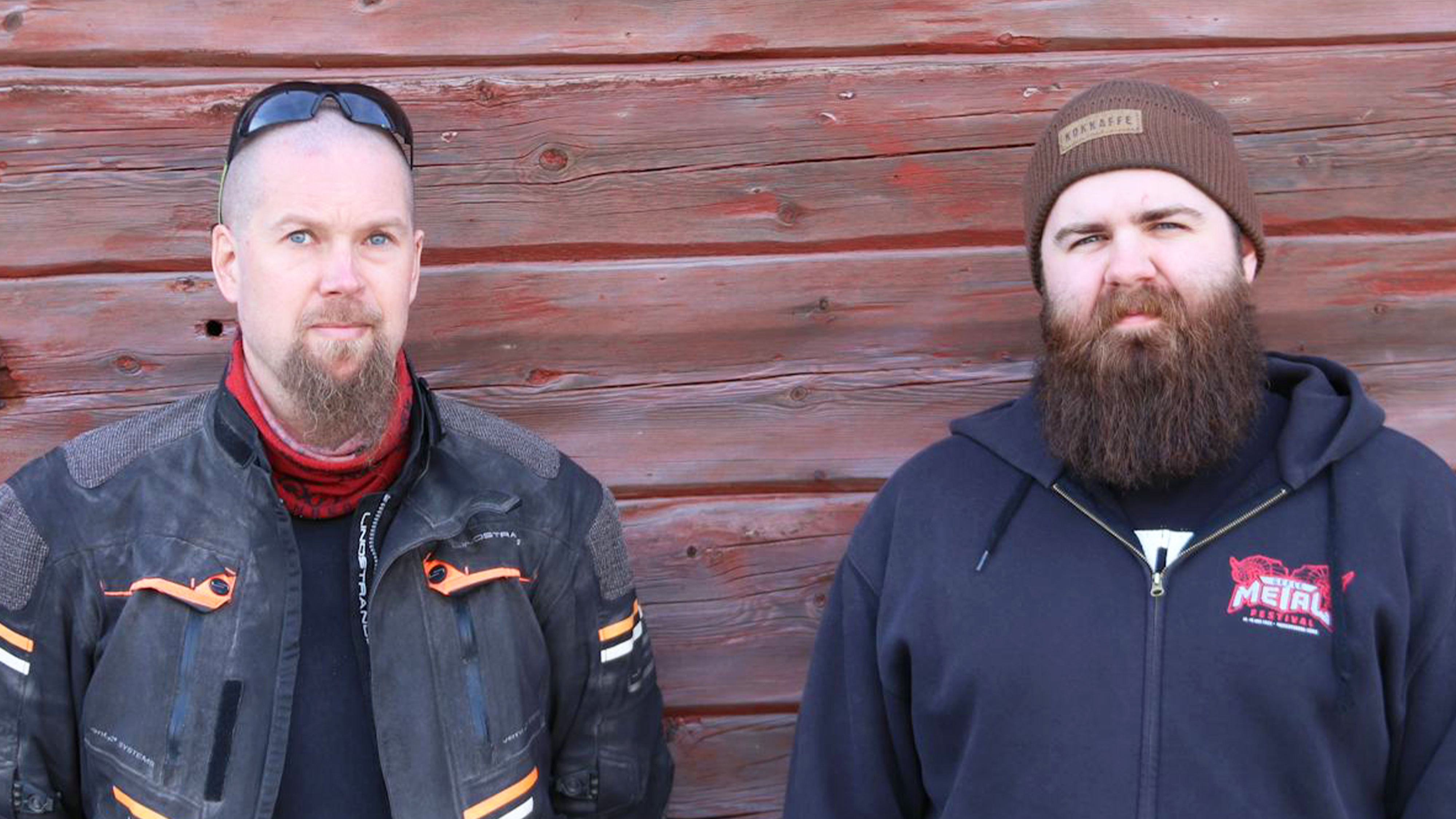 Två män med skägg står framför en trävägg. En har motorcykeljacka och solglasögon på huvudet, den andra mössa och hoodie.