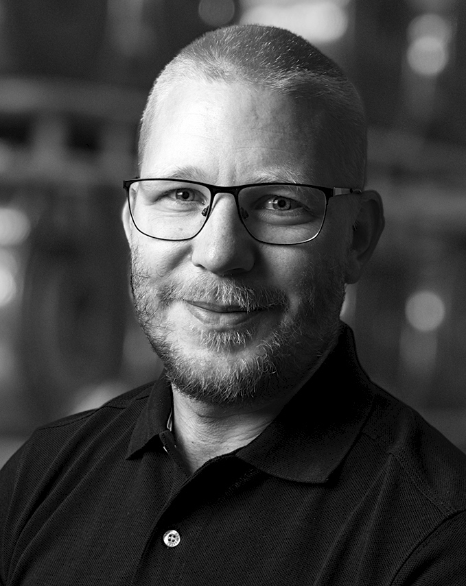 En man med kort hår, glasögon och skägg har på sig en mörk skjorta med krage, tittar mot kameran och ler svagt; suddig bakgrund.