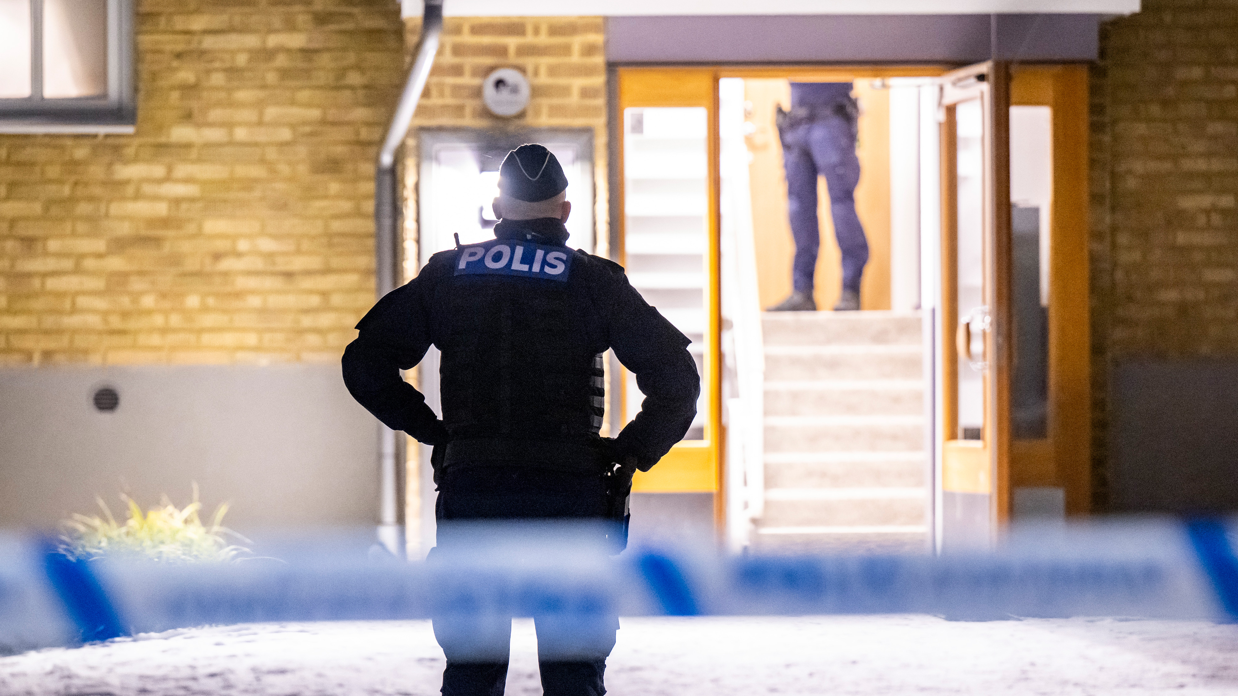 En polis står bakom avspärrningsband utanför en byggnadsentré på natten, och en annan syns inomhus vid trappan.