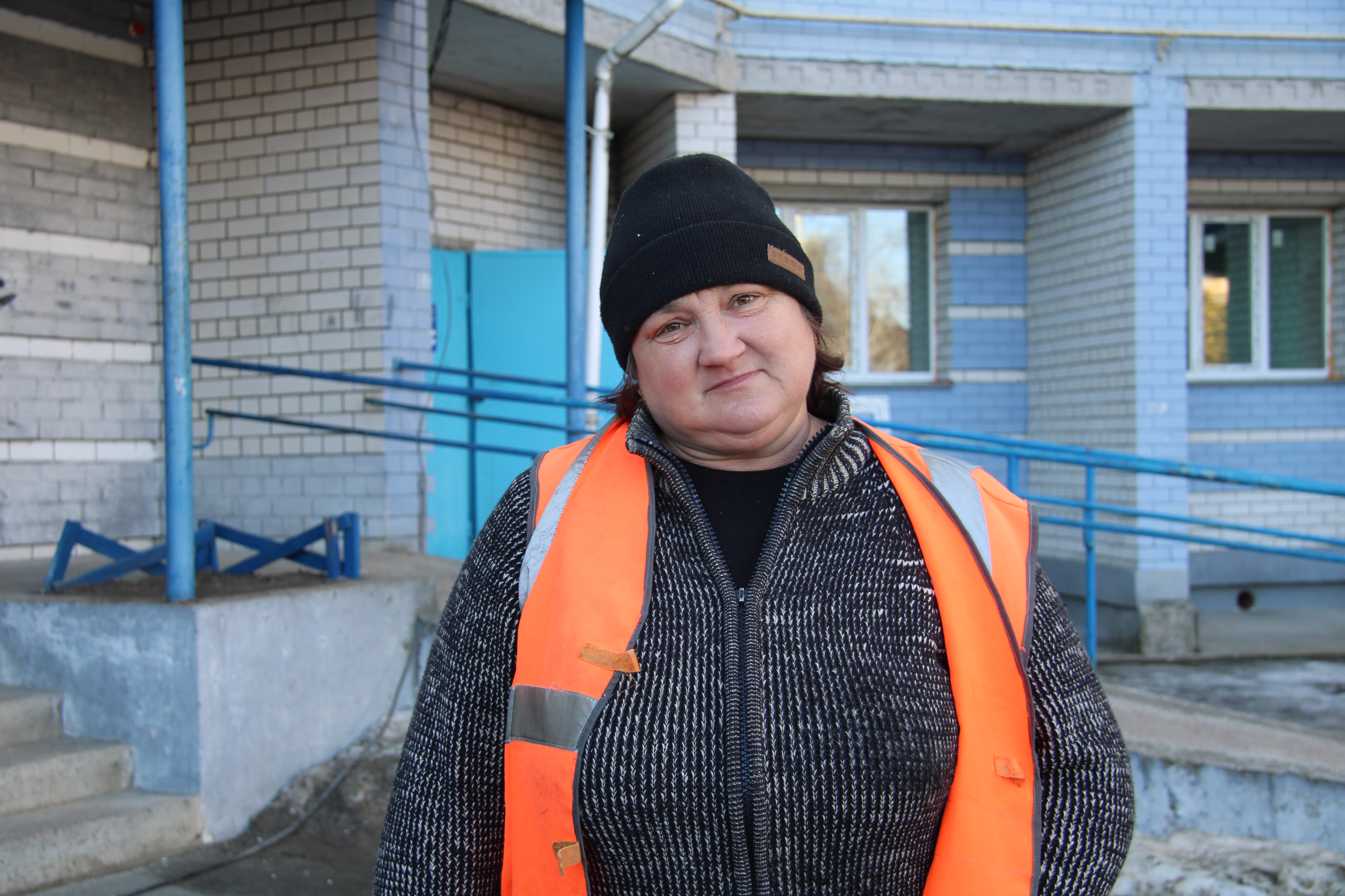 Ludmila Koliadenko med svart mössa, grå tröja med dragkedja och orange reflexväst står framför en tegelbyggnad med blå detaljer.