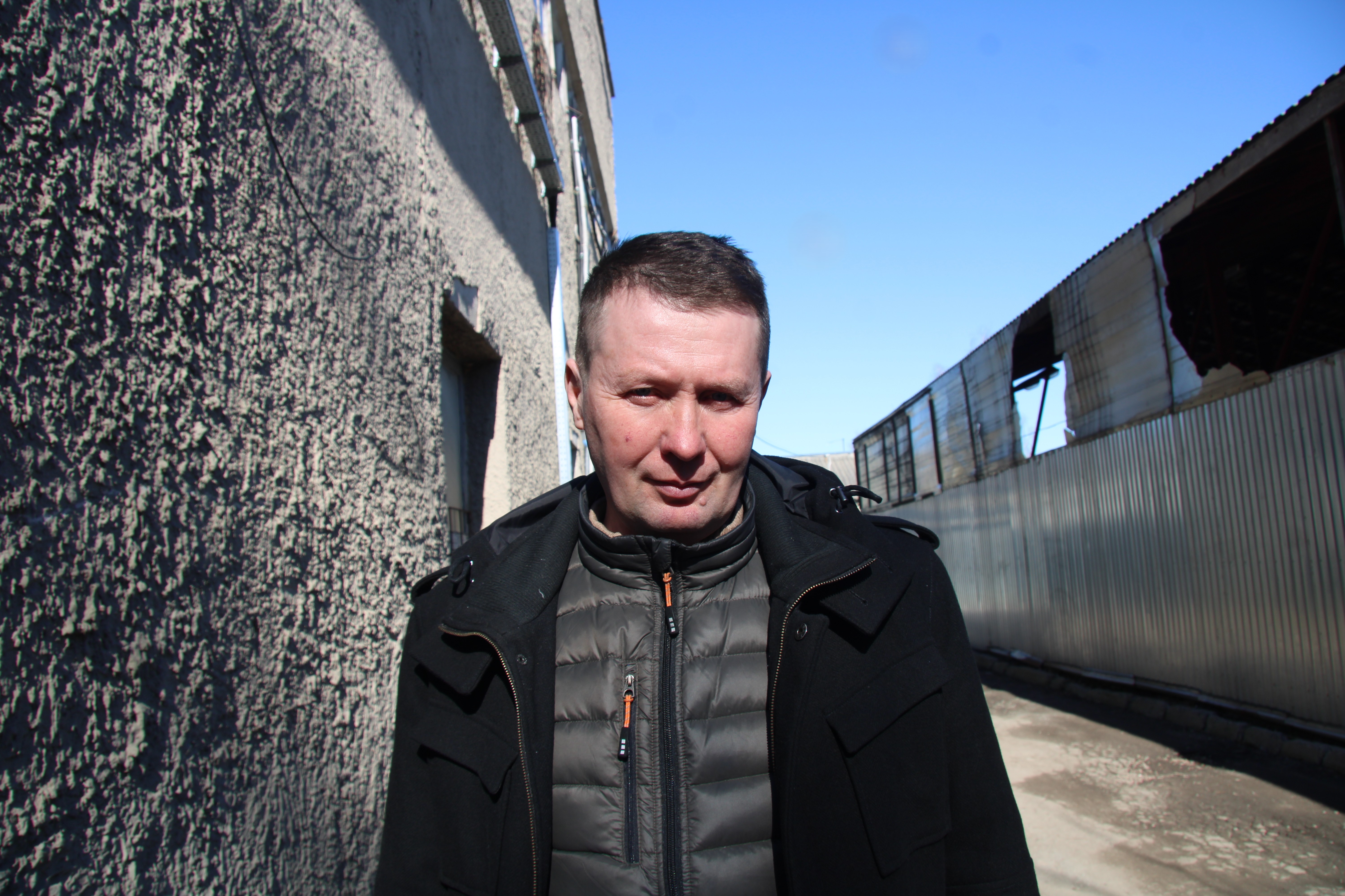 Vasyl Khvostikov står utomhus mellan en vägg och ett korrugerat metallstaket under en blå himmel.