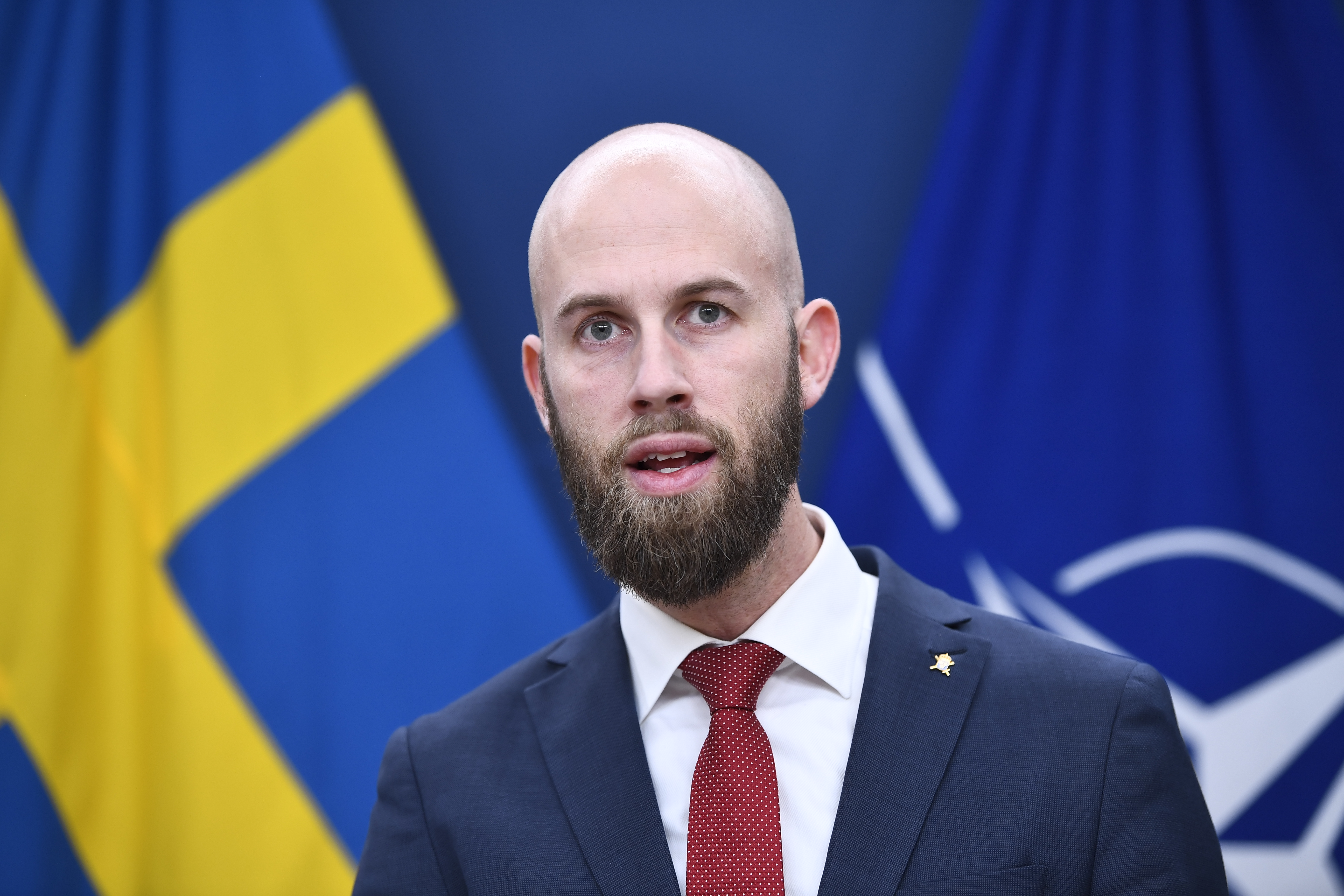 En man i kostym och röd slips talar framför svenska och NATO-flaggor.