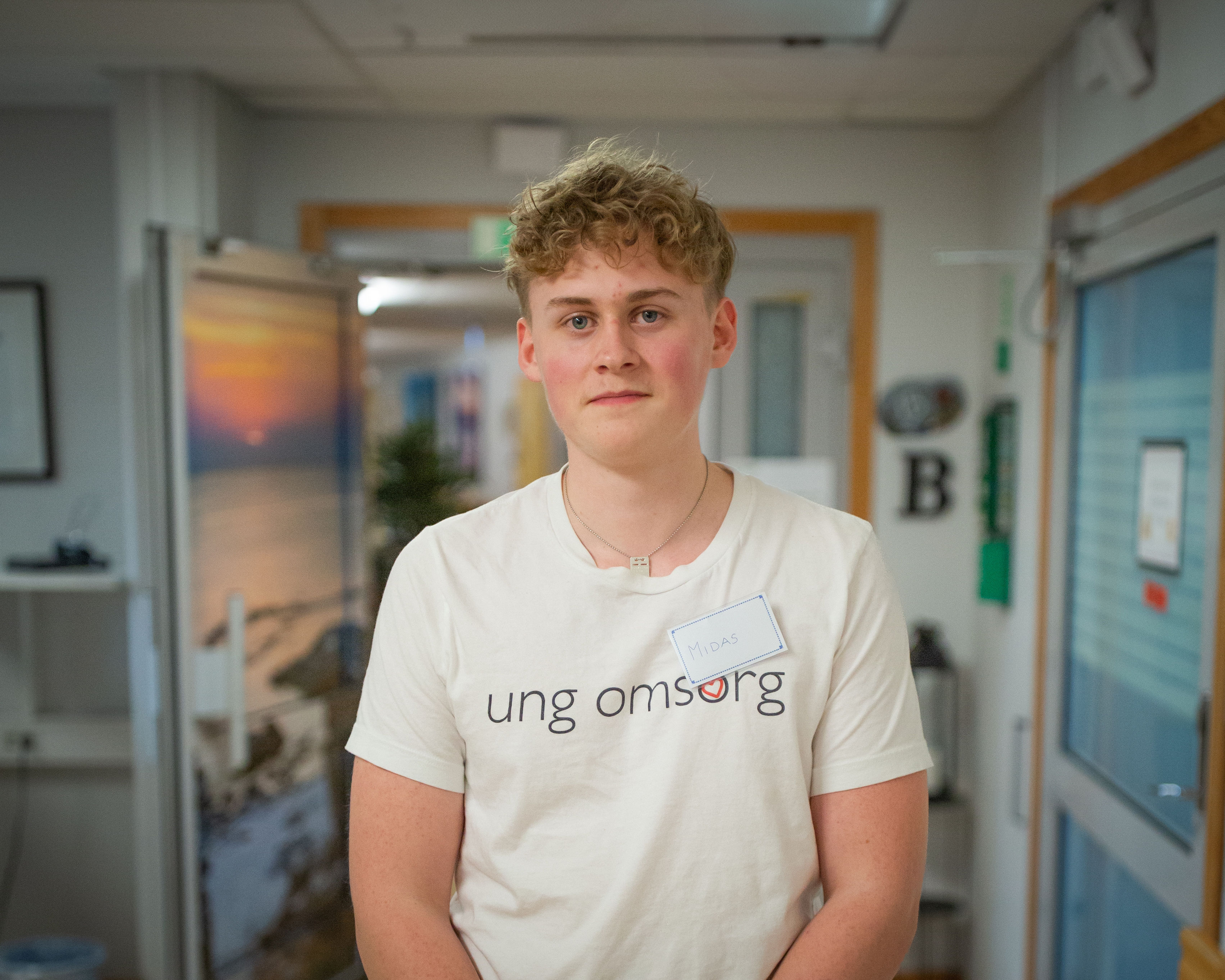En person med kort, lockigt hår står inomhus och har på sig en vit "ung omsorg"-tröja och en namnlapp med texten "Mika".