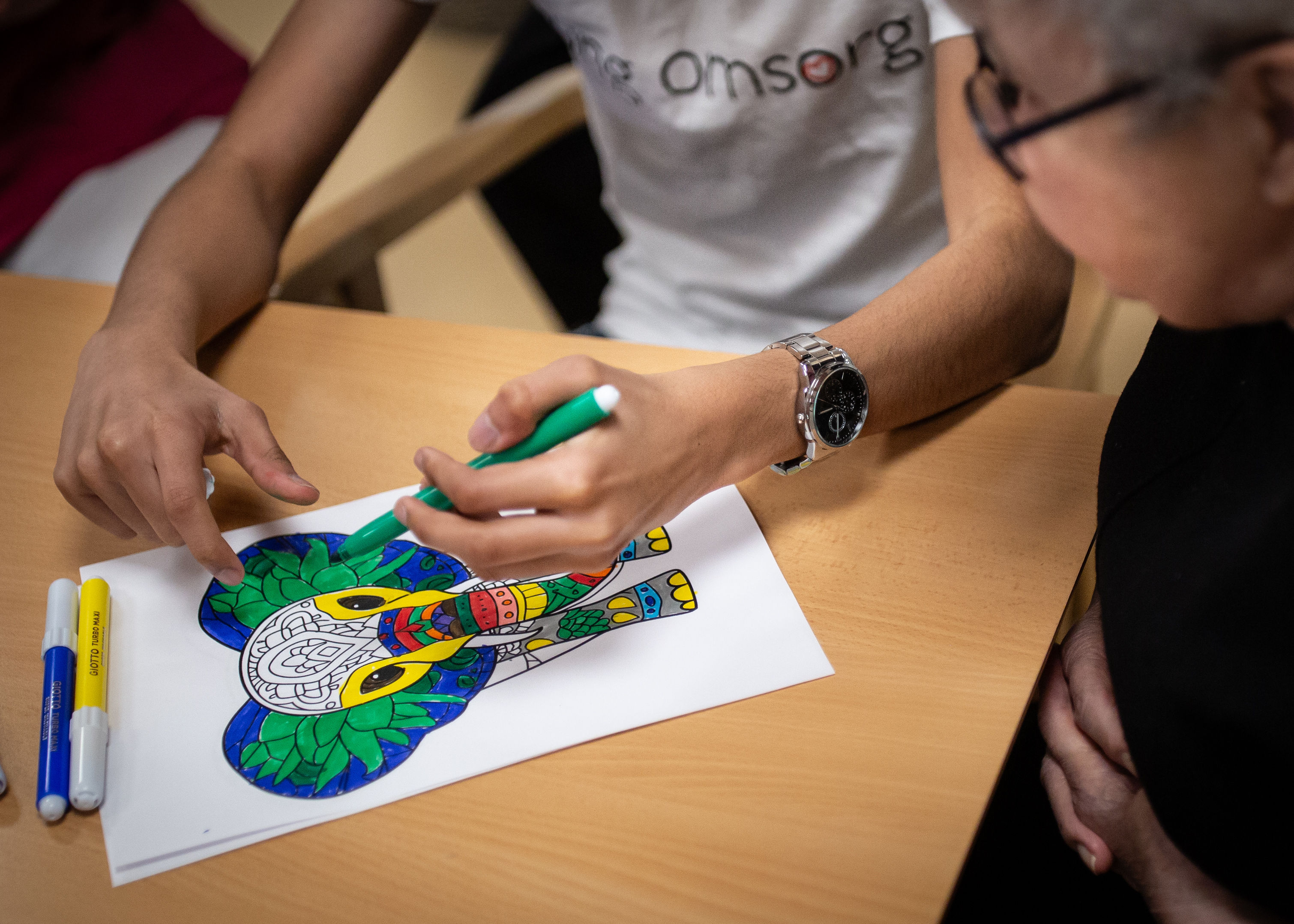 Två personer sitter vid ett bord, en person färgar en bild av en elefant med en tuschpenna och den andra personen tittar på den.