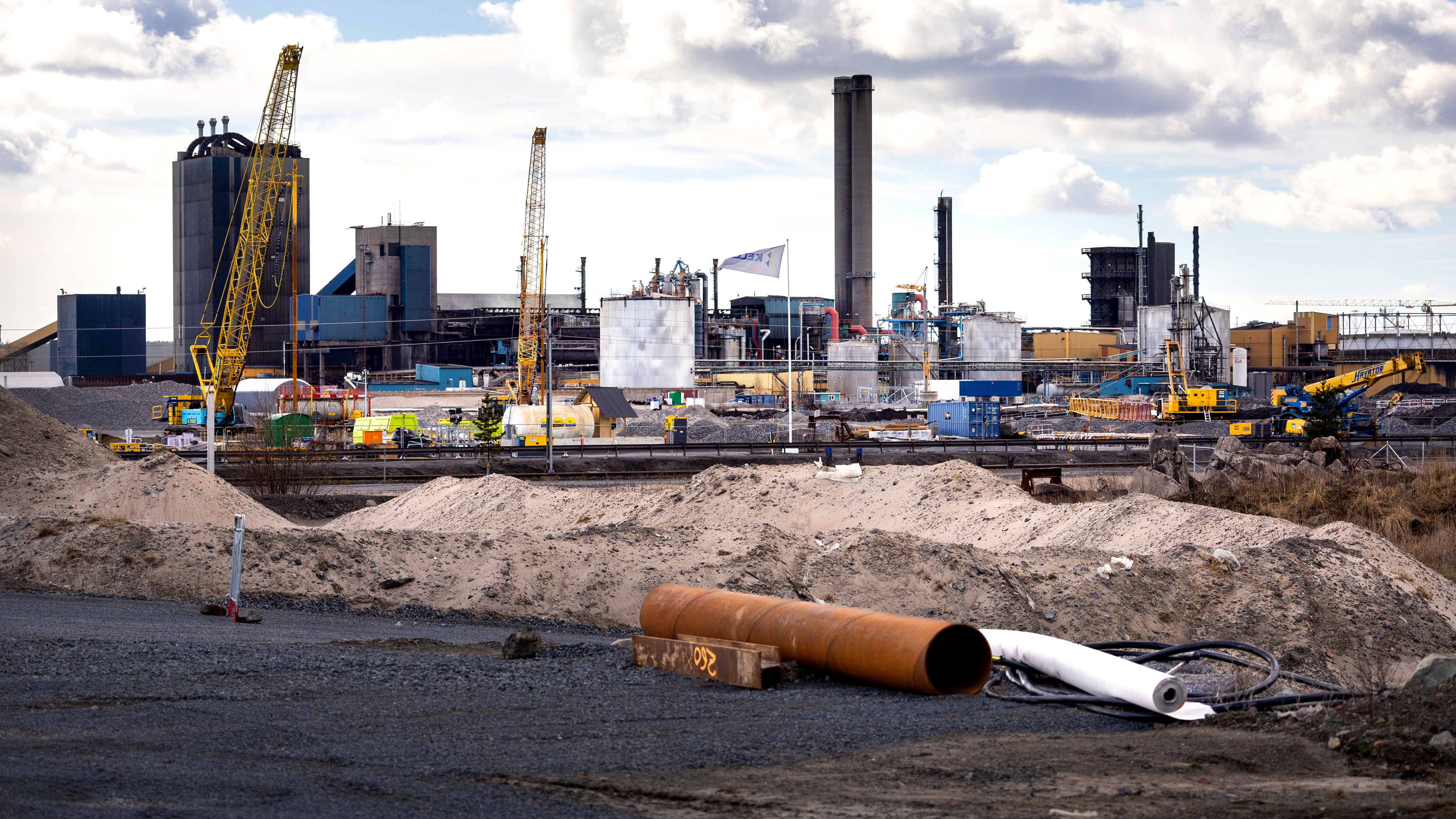 Industriell byggarbetsplats med kranar, maskiner, jordhögar, stora rör och olika fabriksbyggnader under molnig himmel.