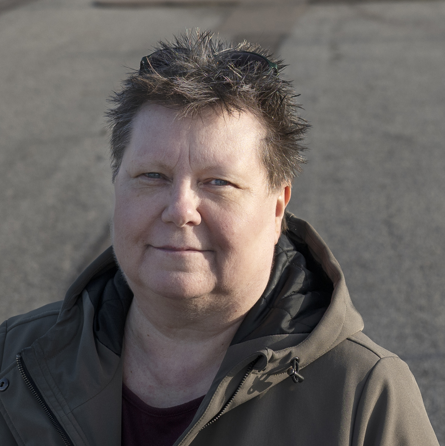 Miriam Håkansson, med kort, ovårdat hår, står utanför på den asfalterade marken, klädd i grön jacka och mörk tröja och stirrar in i kameran.