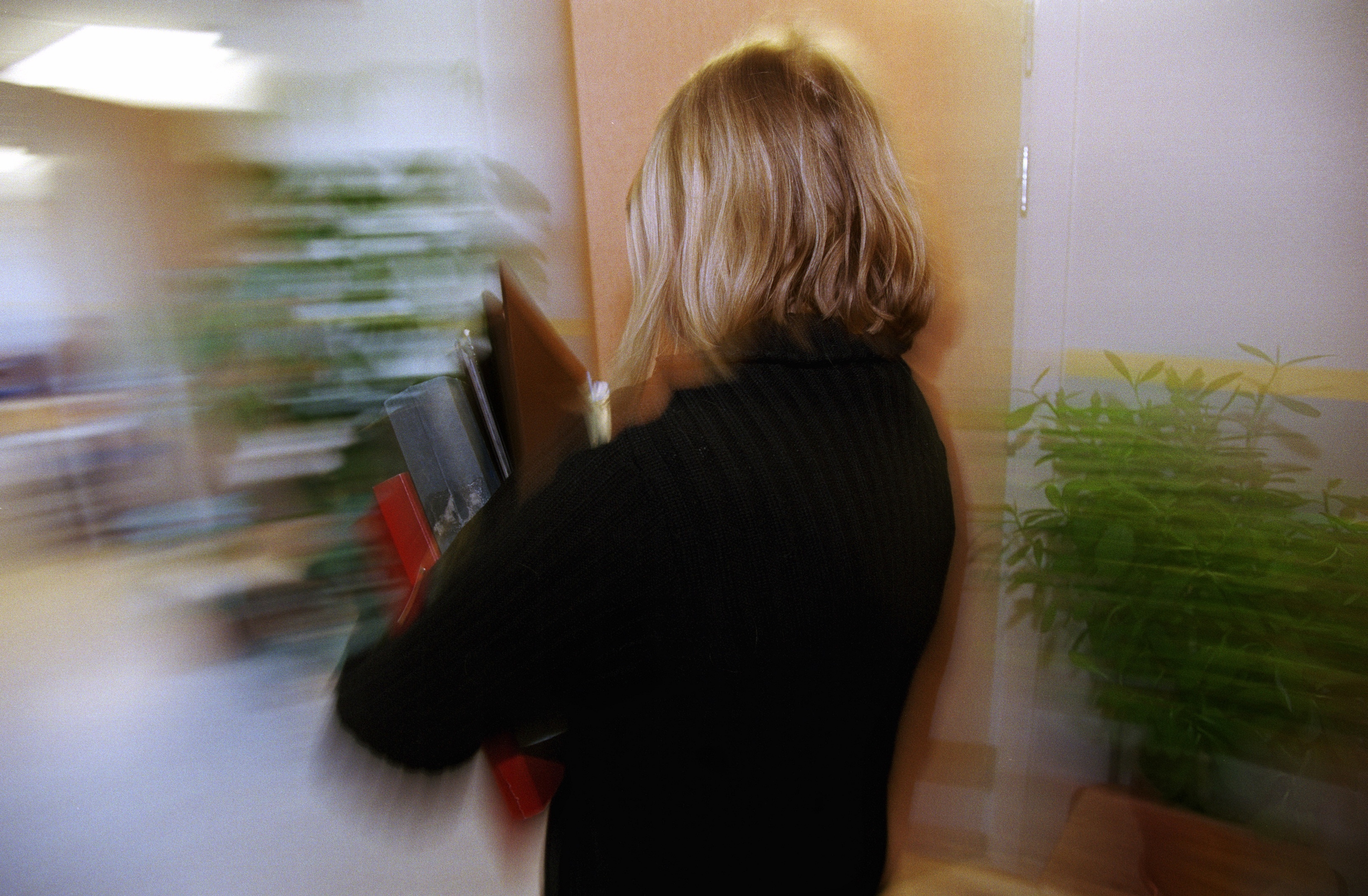 En person med blont hår och svart tröja bär på pärmar och går inomhus; bilden är suddig på grund av rörelse.