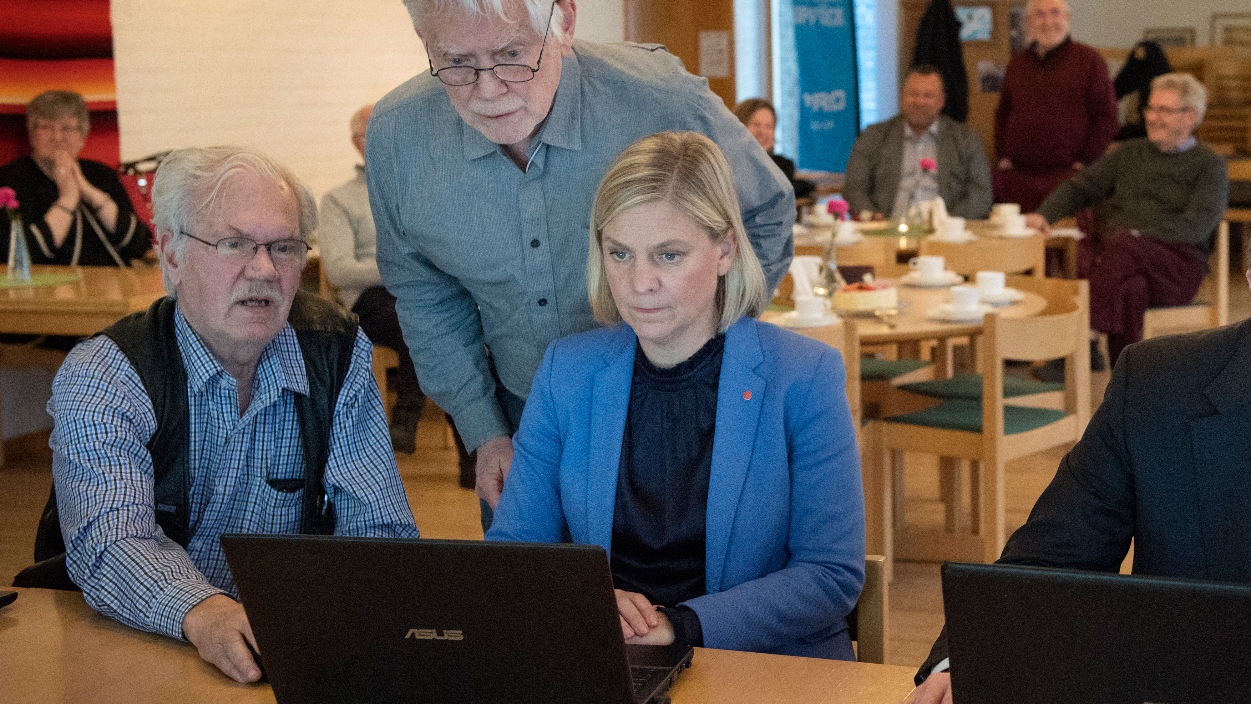 En kvinna i blå kavaj sitter vid en bärbar dator mellan två äldre män i ett rum där flera personer sitter vid bord.