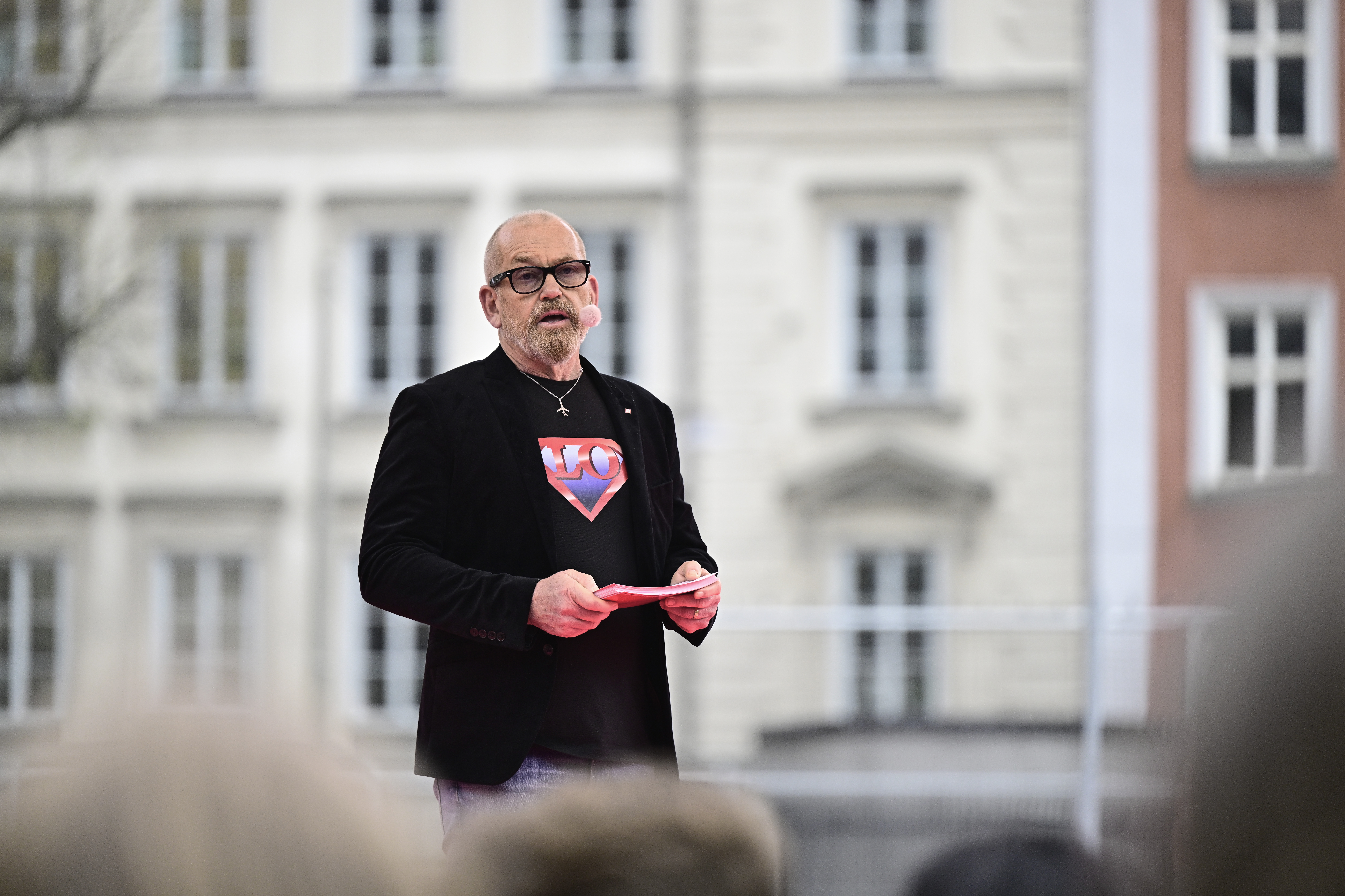 En man med glasögon och skägg, klädd i en svart kavaj över en Superman-tröja, pratar utomhus med ett papper i handen. En byggnad kan ses i bakgrunden.