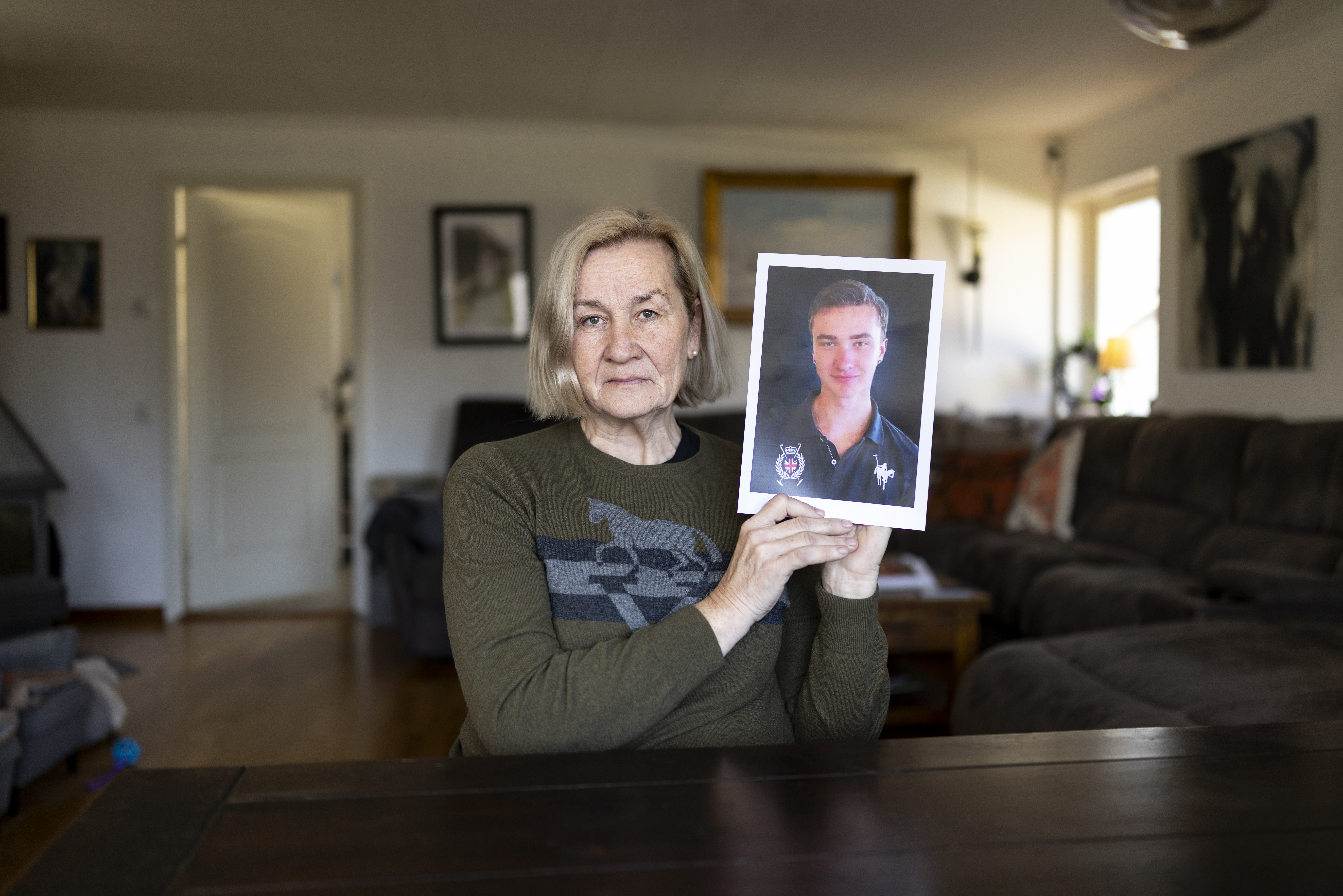 En äldre kvinna sitter vid ett bord i ett vardagsrum, håller upp ett utskrivet foto av en ung man och tittar mot kameran.