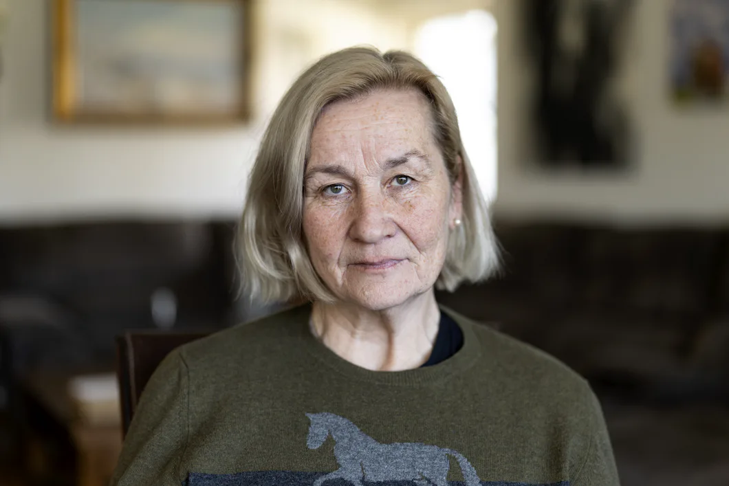 En äldre kvinna med kort blont hår och androgynt ansikte sitter inomhus iförd en grön tröja med hästmotiv. Bakgrunden är suddig.