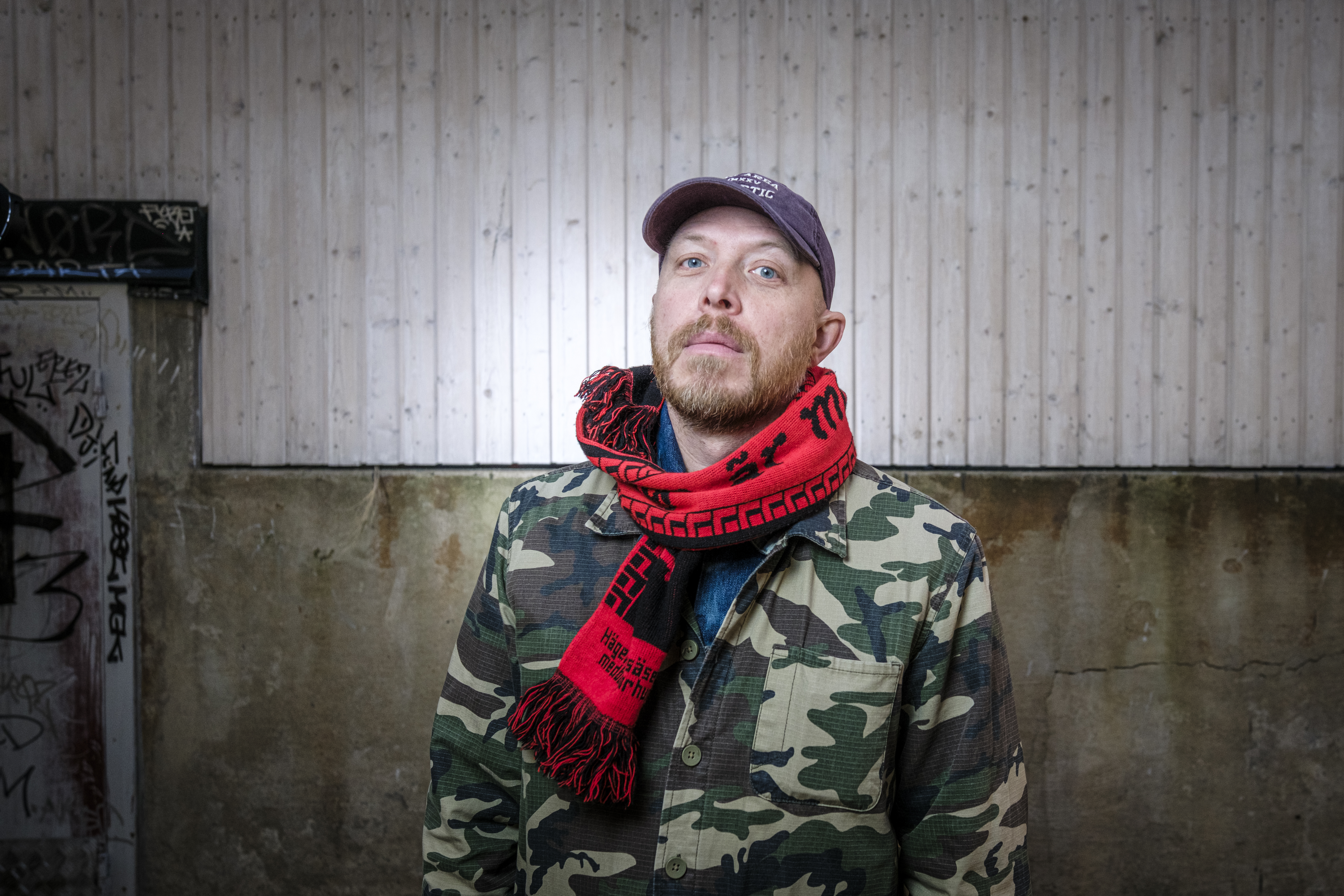 En man med kamouflagejacka, röd och svart halsduk samt mörk keps står framför en betongvägg med graffiti.