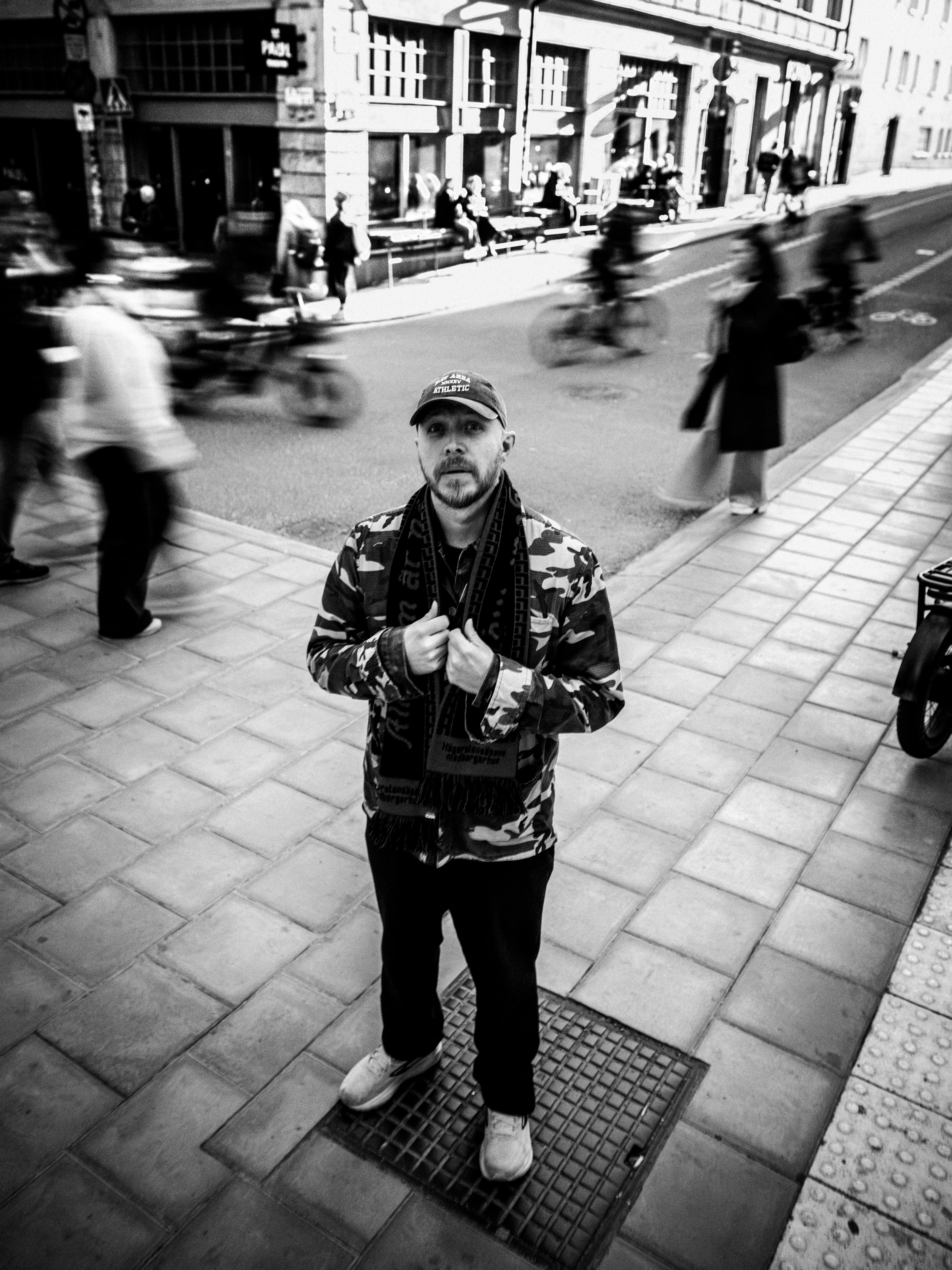 En man med kamouflagejacka står på en trottoar i staden medan fotgängare och cyklister rör sig sig på gatan bakom honom.