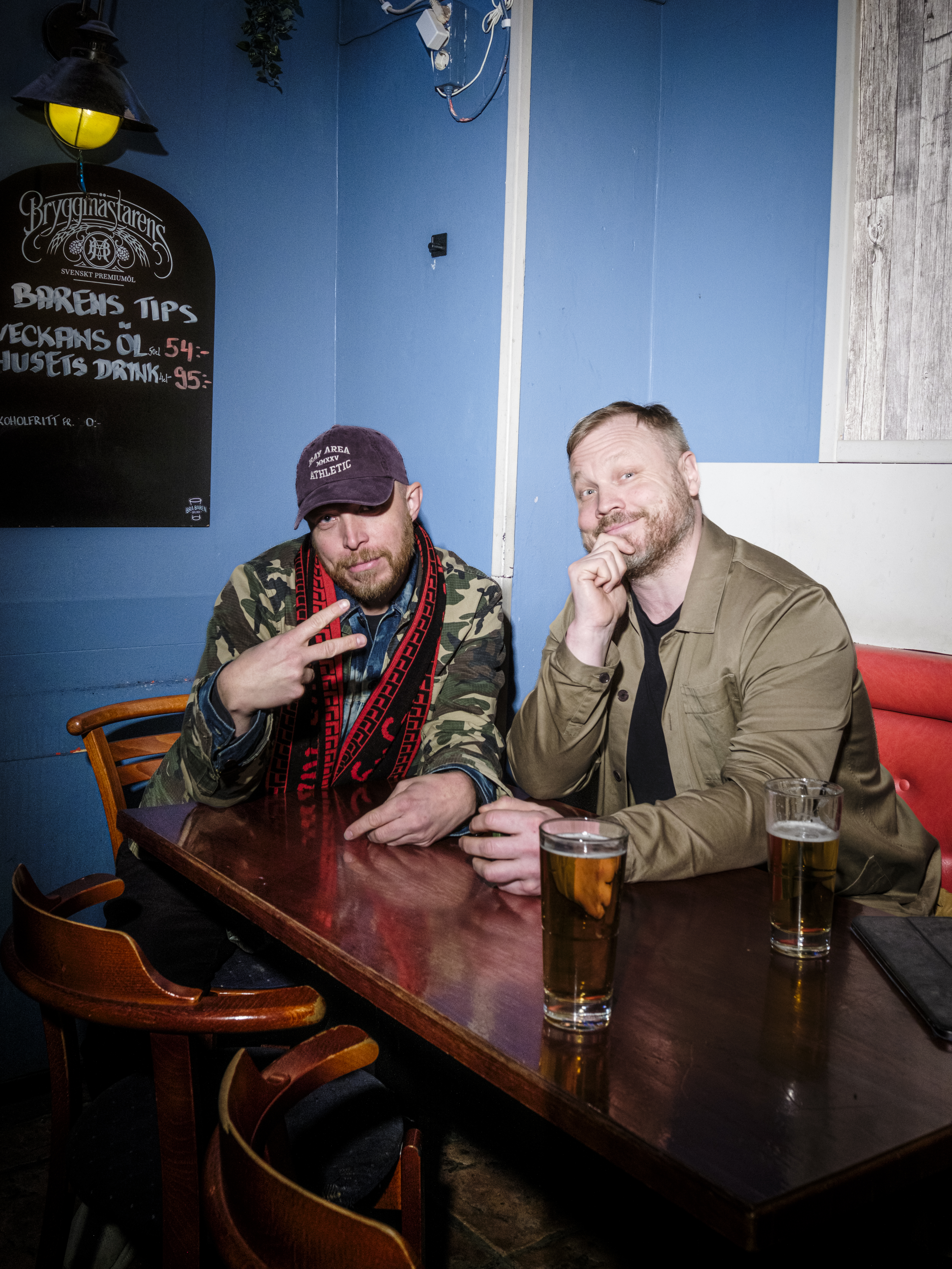 Två män sitter vid ett barbord med drinkar framför sig. En person gör ett fredstecken och den andra lägger handen på hakan.