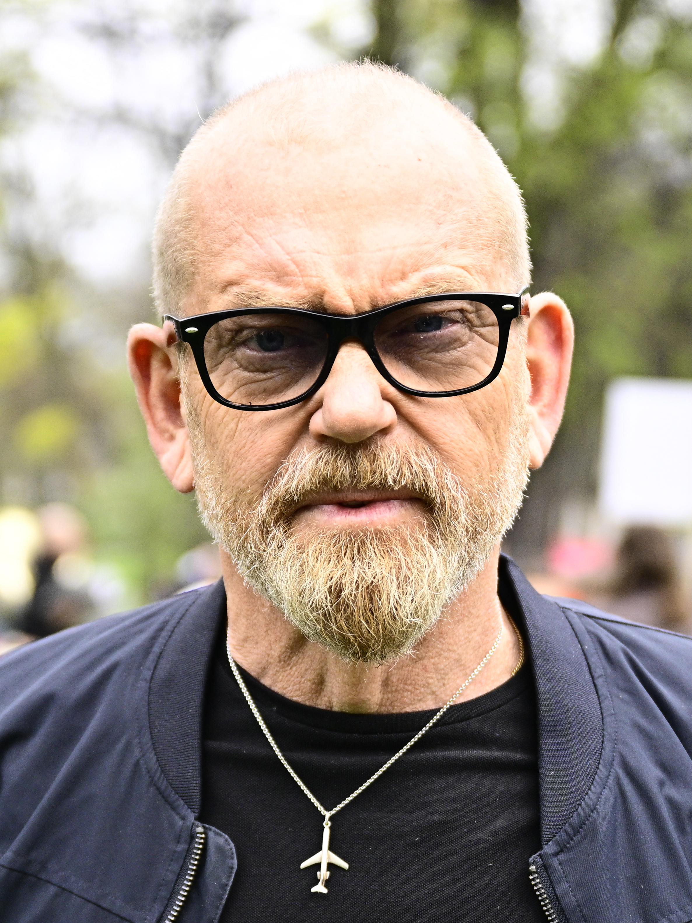 Johan Lindholm, med rakat huvud och skägg, bär glasögon, en svart skjorta, en svart jacka och ett flygplanshalsband. Han står utanför bland träden.