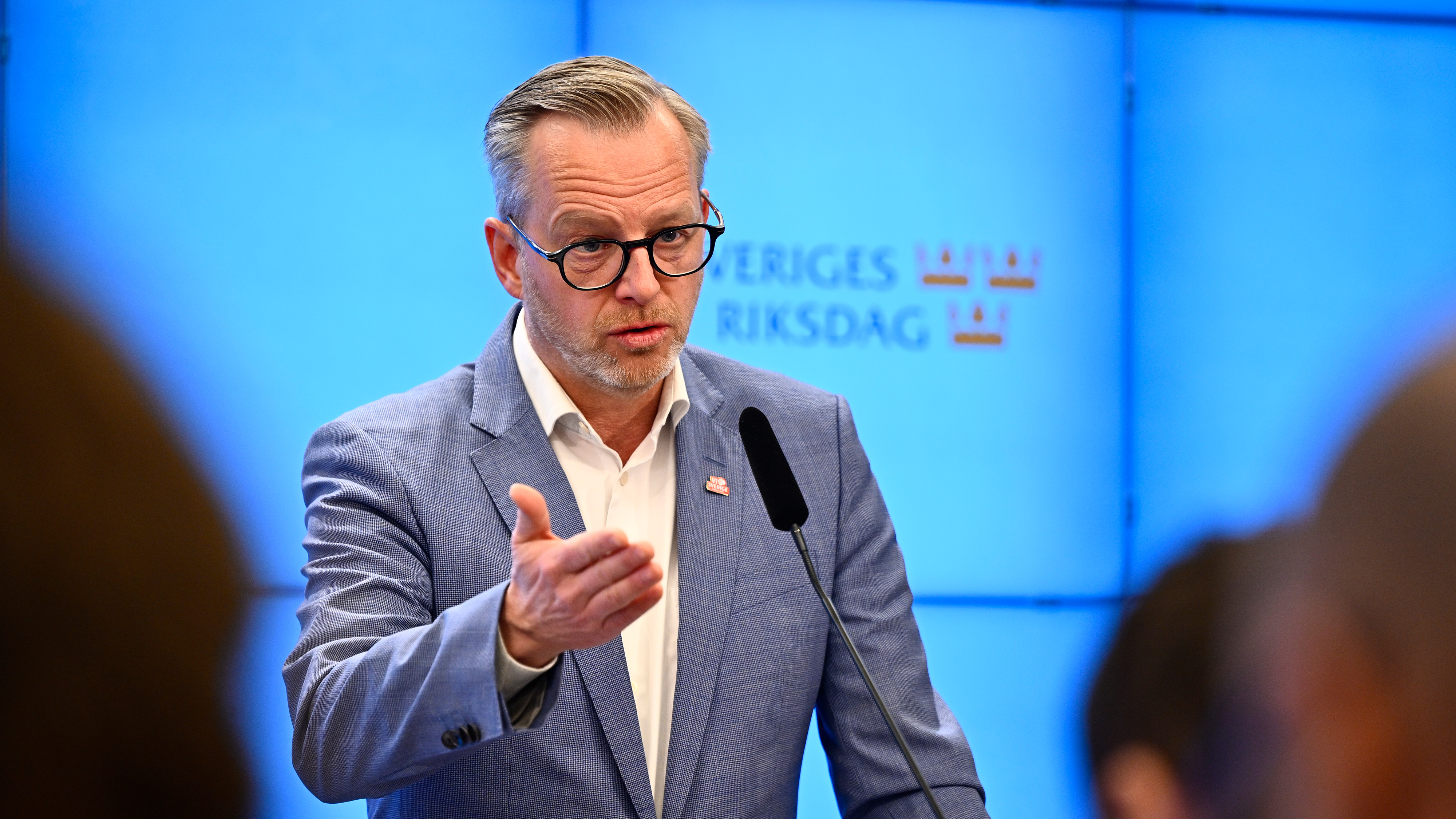 En man i blå kostym och glasögon talar vid ett podium med mikrofon framför en blå bakgrund med texten "Sveriges Riksdag" och kronor.