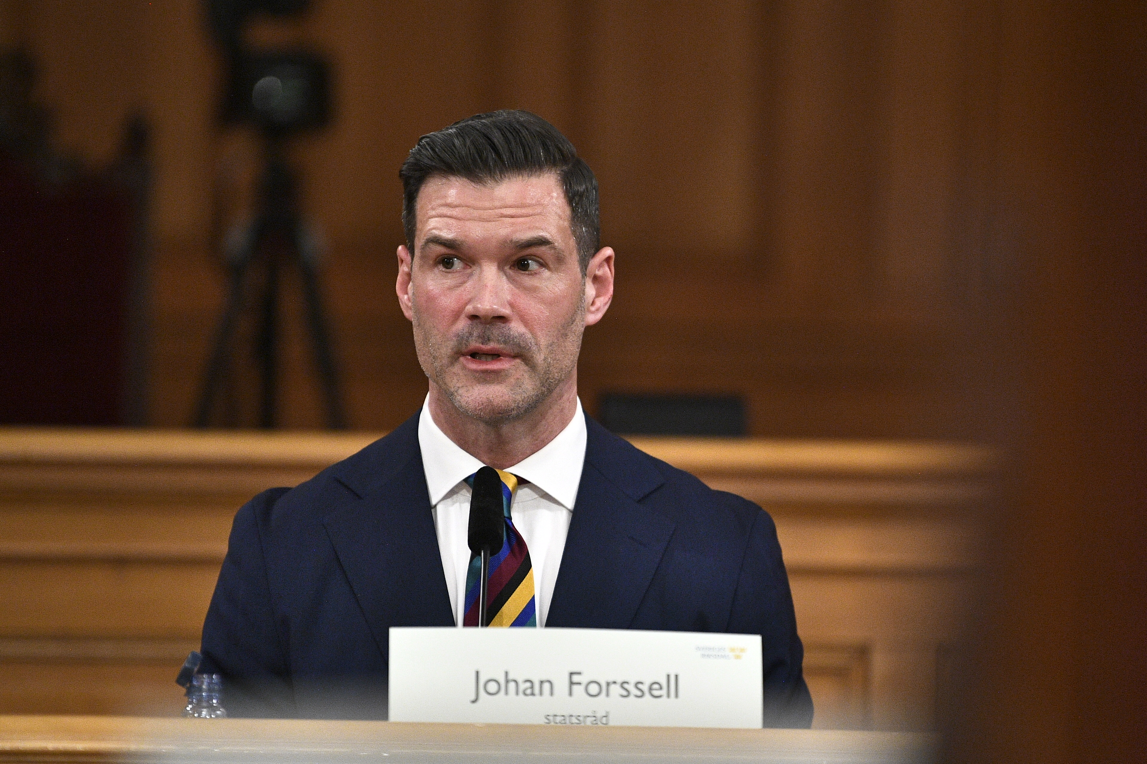 En man i kostym och randig slips talar vid ett podie med en namnskylt där det står "Johan Forssell" inomhus.