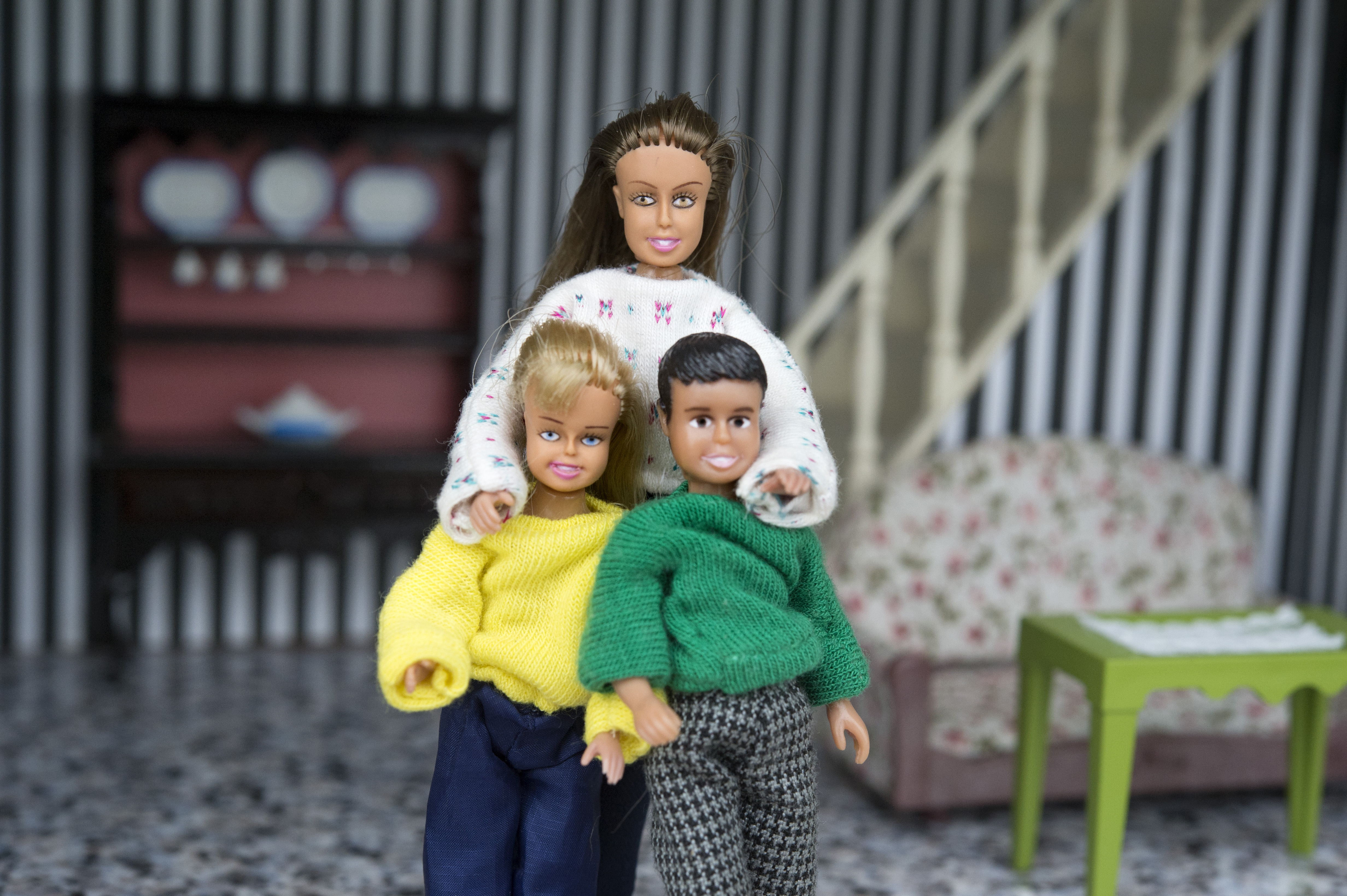 Tre dockor är placerade i ett dockskåps vardagsrum; en vuxendocka står bakom två barndockor, alla har på sig tröjor.