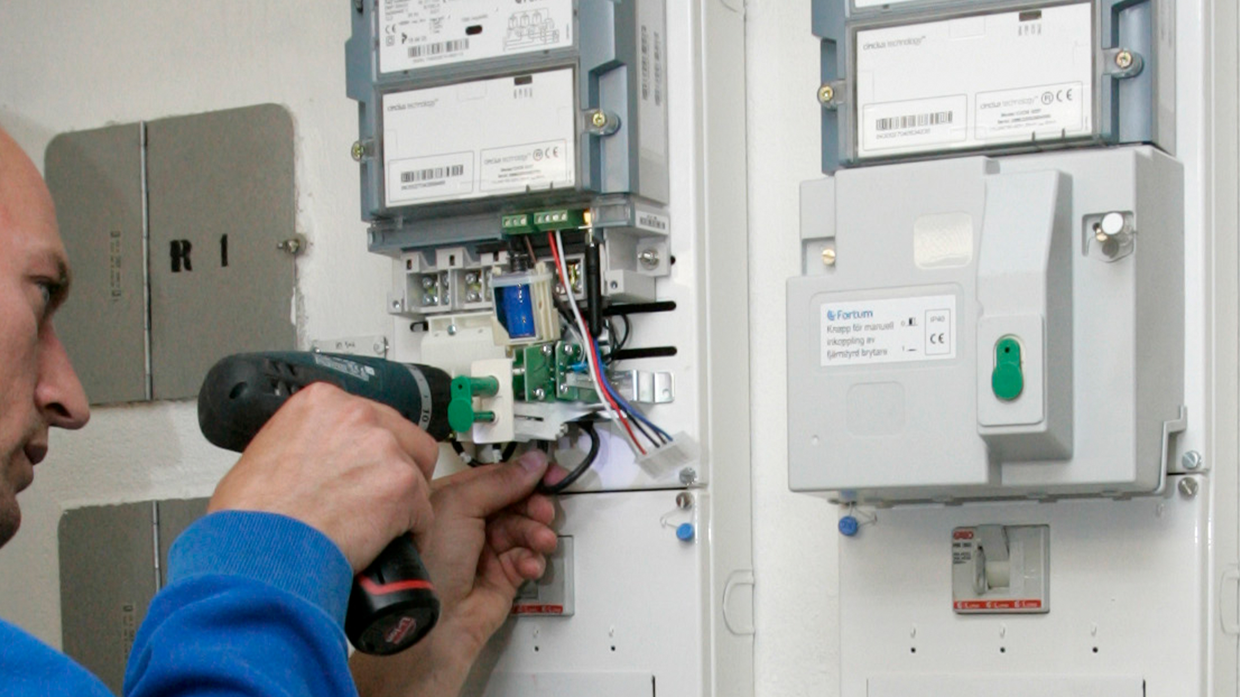 En person använder en borrmaskin för att utföra underhåll eller installation på en elmätarpanel med exponerade ledningar.