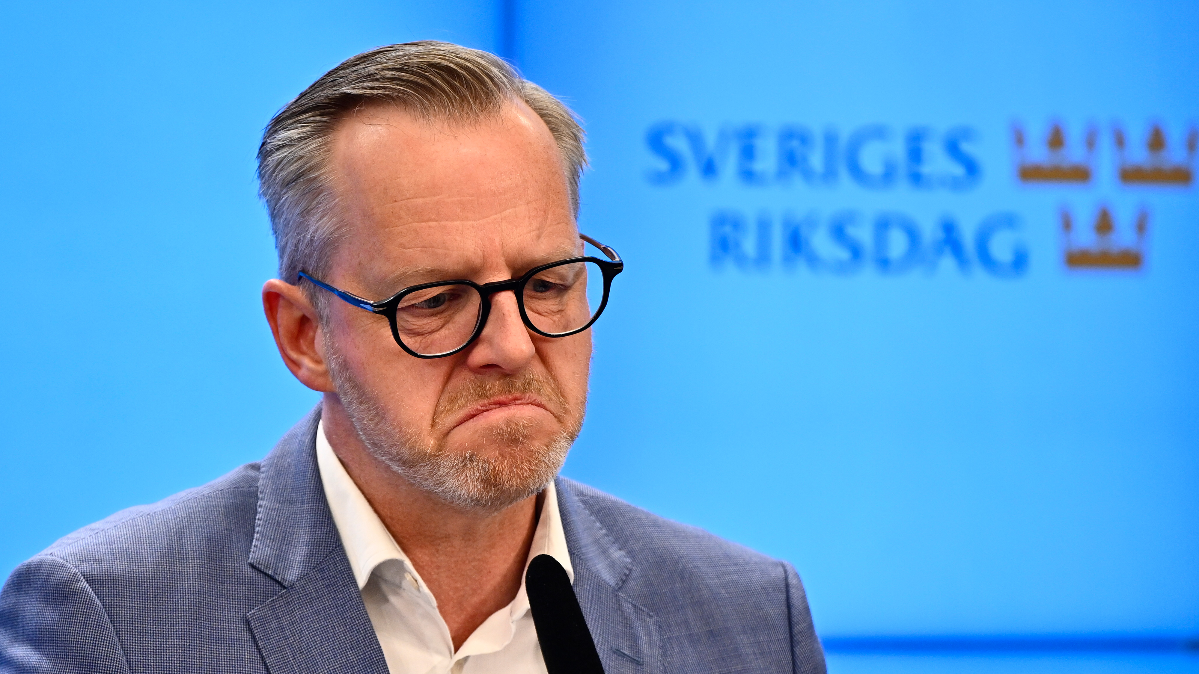 Mikael Danberg, iklädd glasögon och kostym, står framför en skylt där det står ``Sveriges riksdag'' och tittar ner.