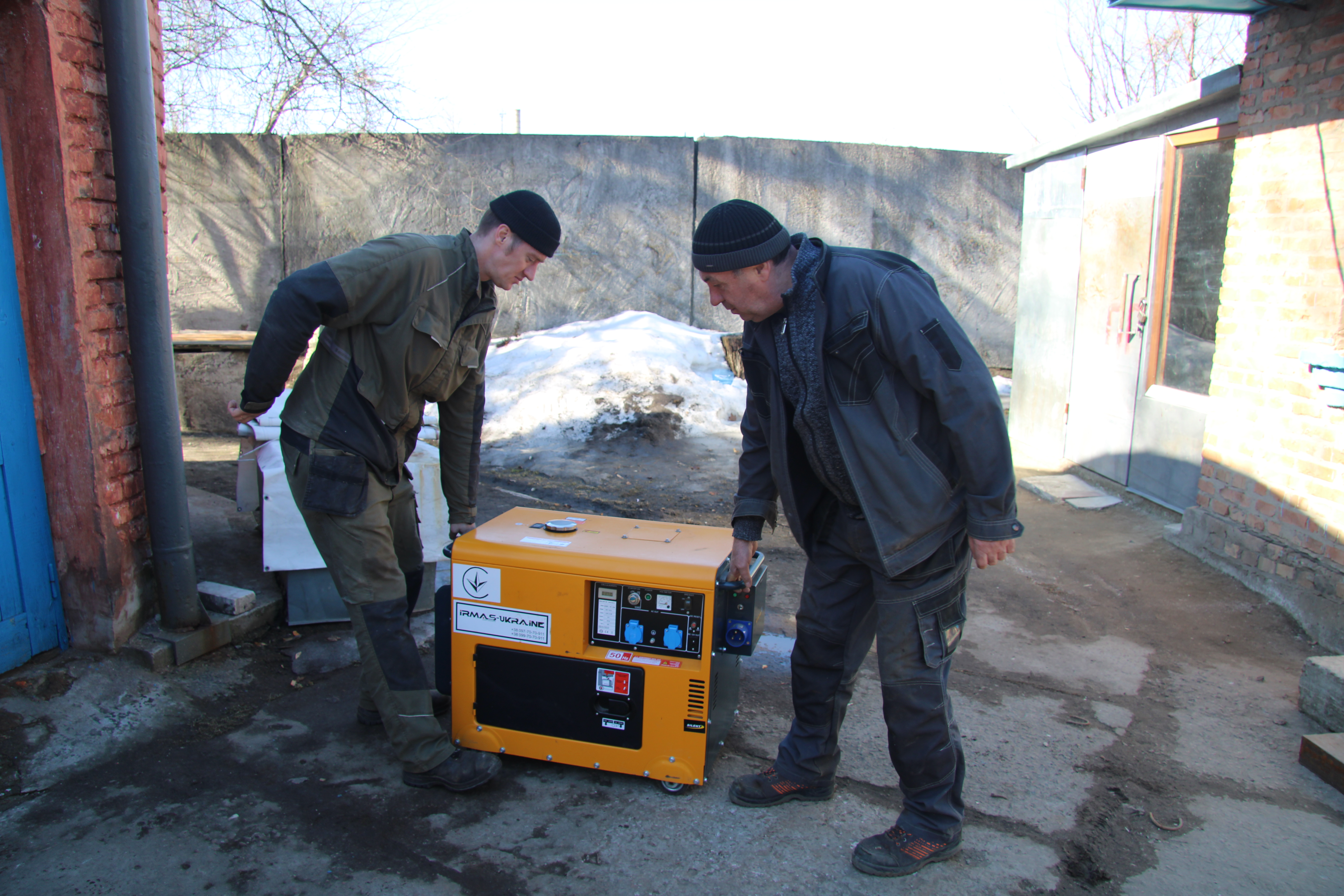 Oleksandr Vorobyov och Valery Shkaro, klädda i arbetskläder, står utanför med en gul bärbar generator på asfalt och smältande snö nära en tegelvägg.