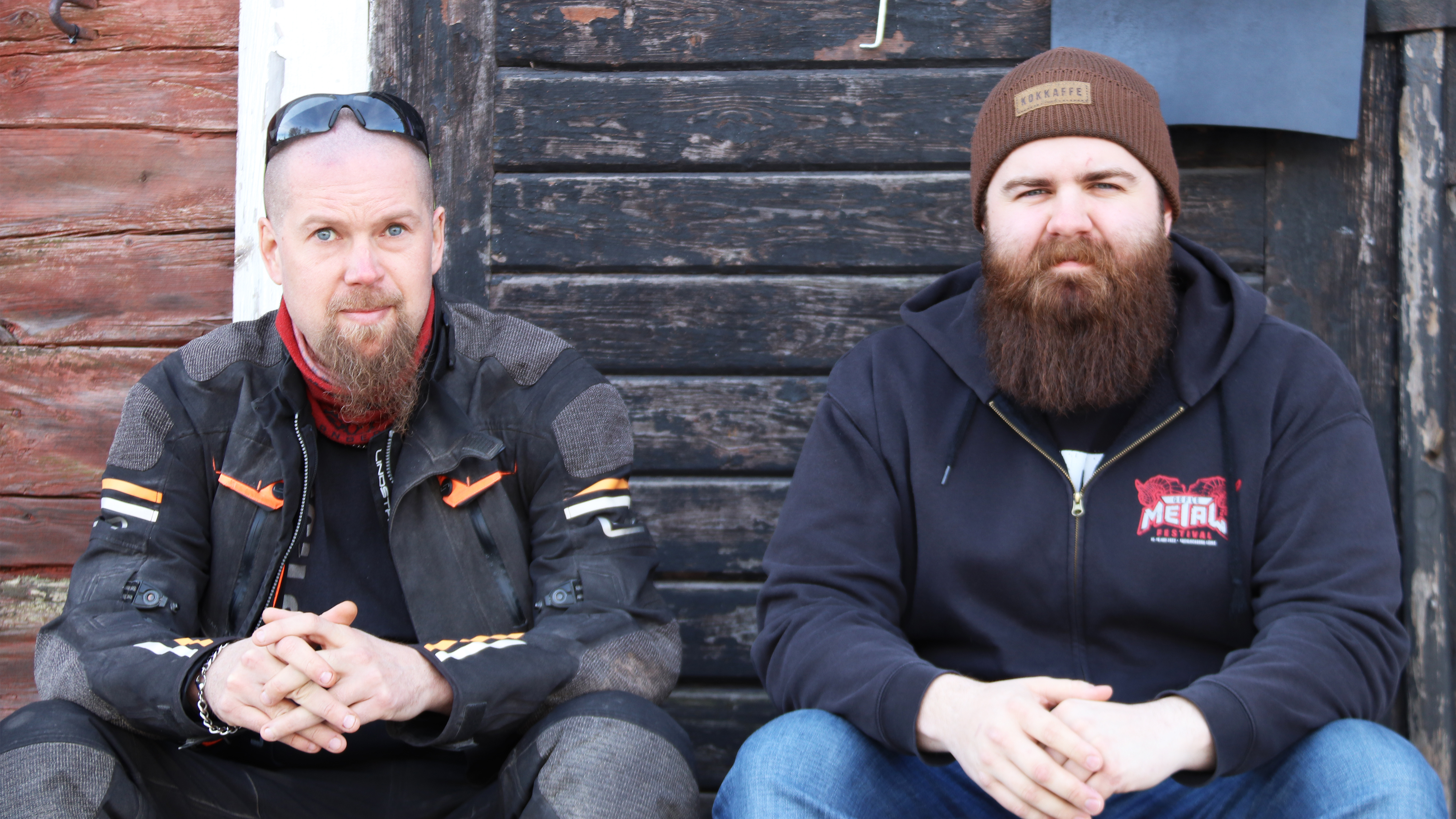 Två män sitter sida vid sida framför en mörk trädörr. Den ene har motorcykeljacka och solglasögon på huvudet och den andre har brun hatt och luvtröja.
