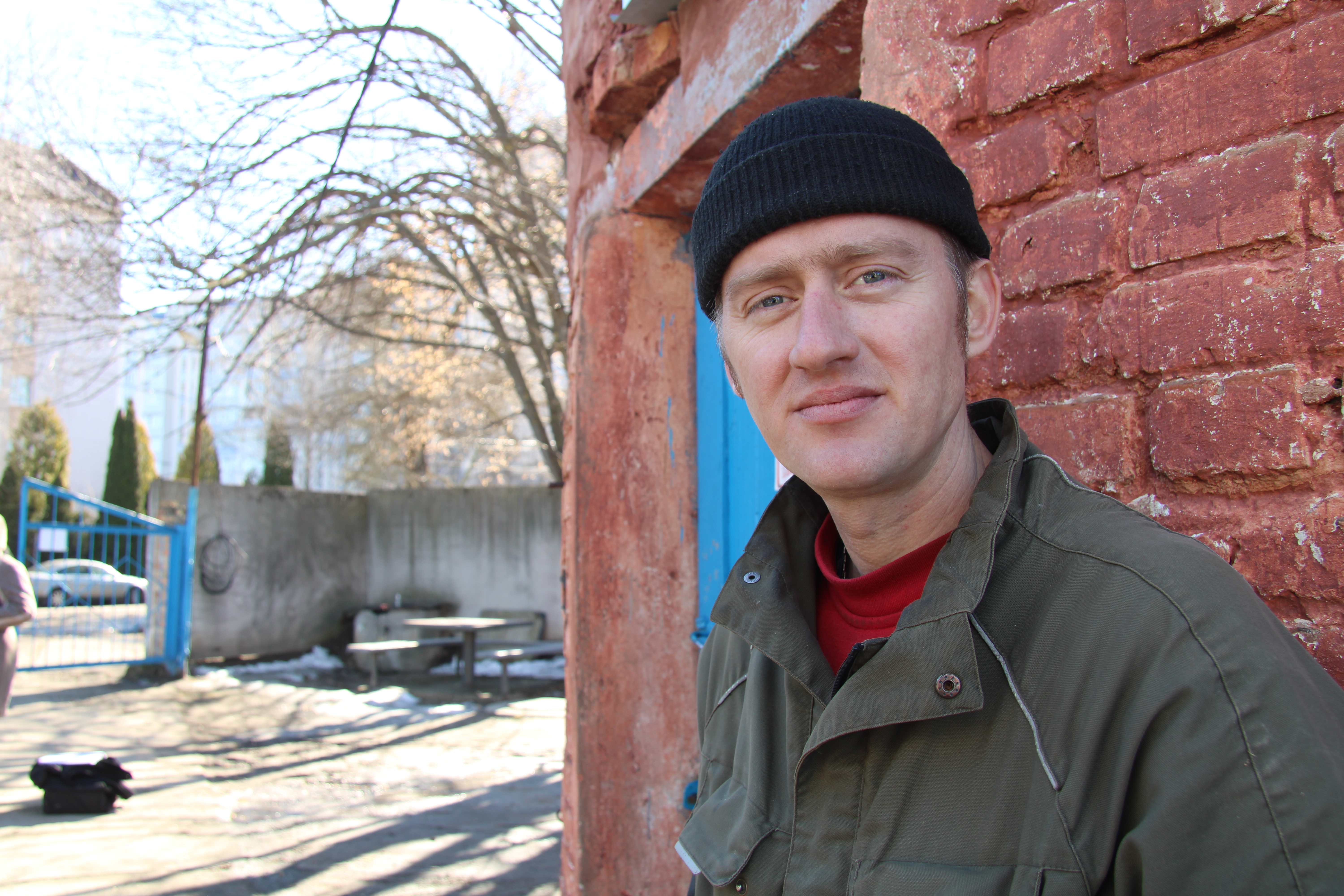 En solig dag står Oleksandr Vorobyov i svart hatt och grön jacka vid en röd tegelvägg.