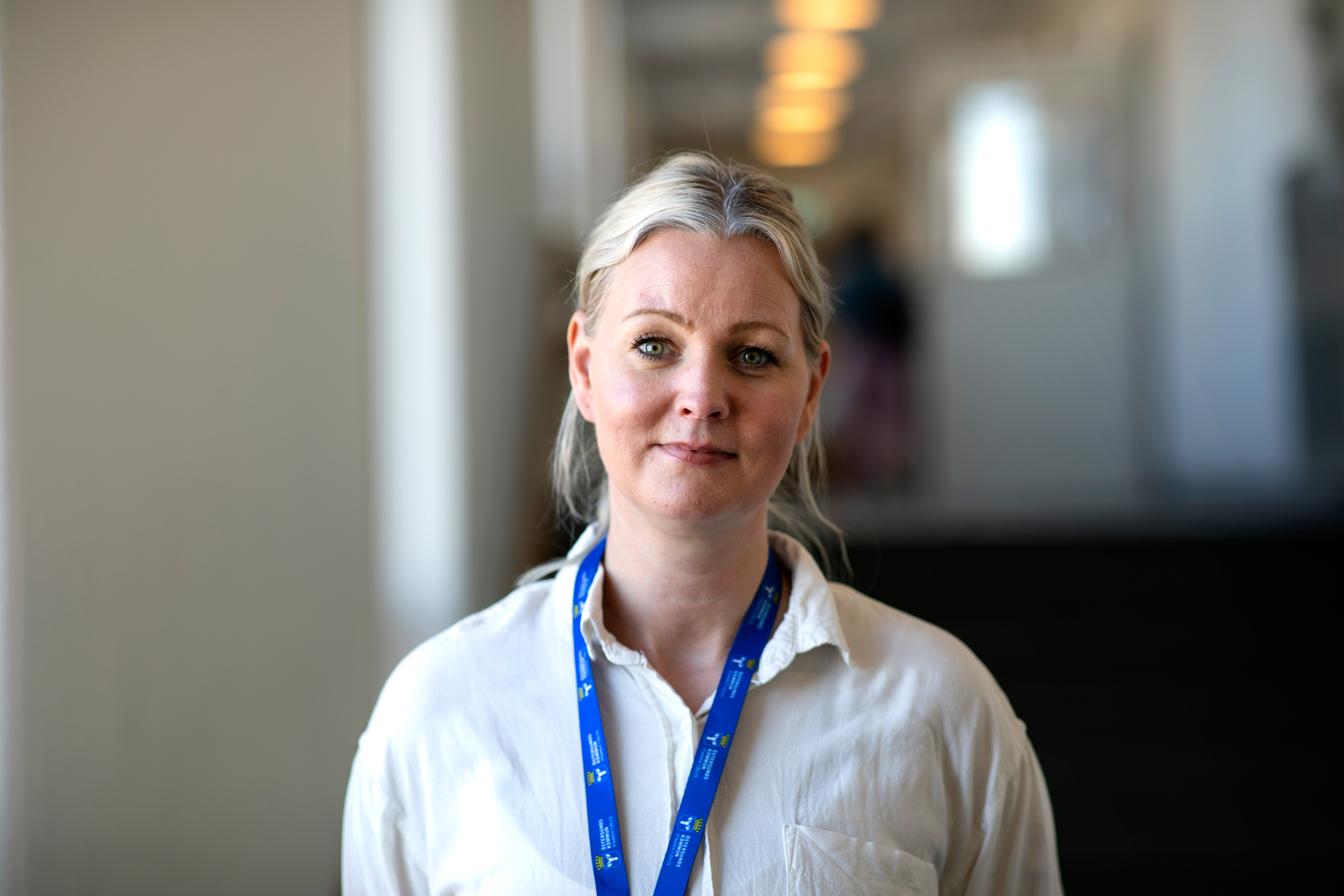 En kvinna med blont hår uppsatt, iklädd vit skjorta och blåa remmar, står i en hall inomhus.