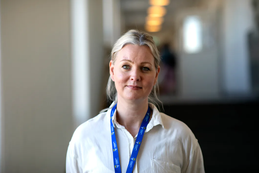 En kvinna med blont hår uppsatt, iklädd vit skjorta och blåa remmar, står i en hall inomhus.