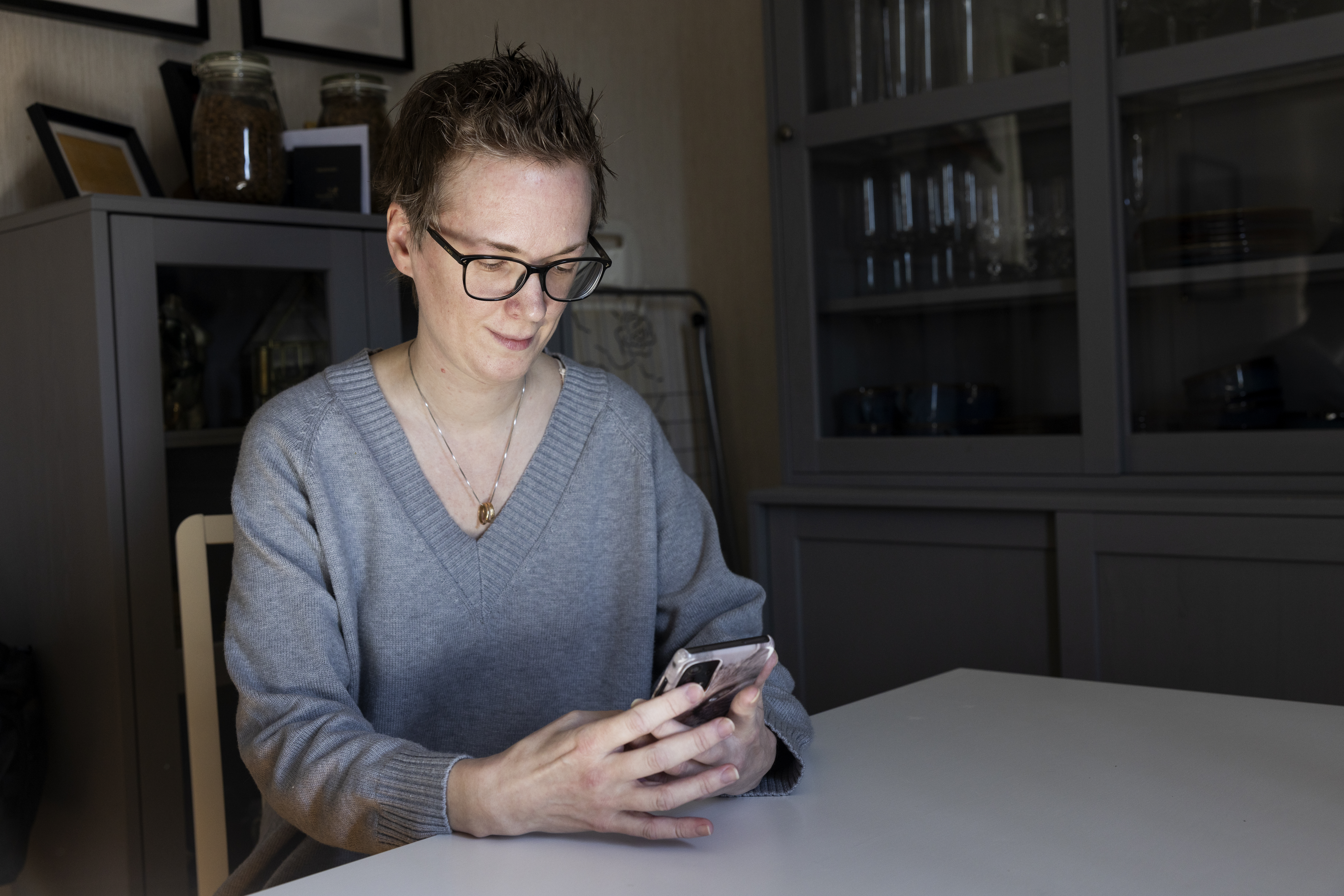 En person med kort hår och glasögon sitter vid ett vitt bord och tittar på en mobiltelefon. I bakgrunden syns hyllor med flaskor och keramik.