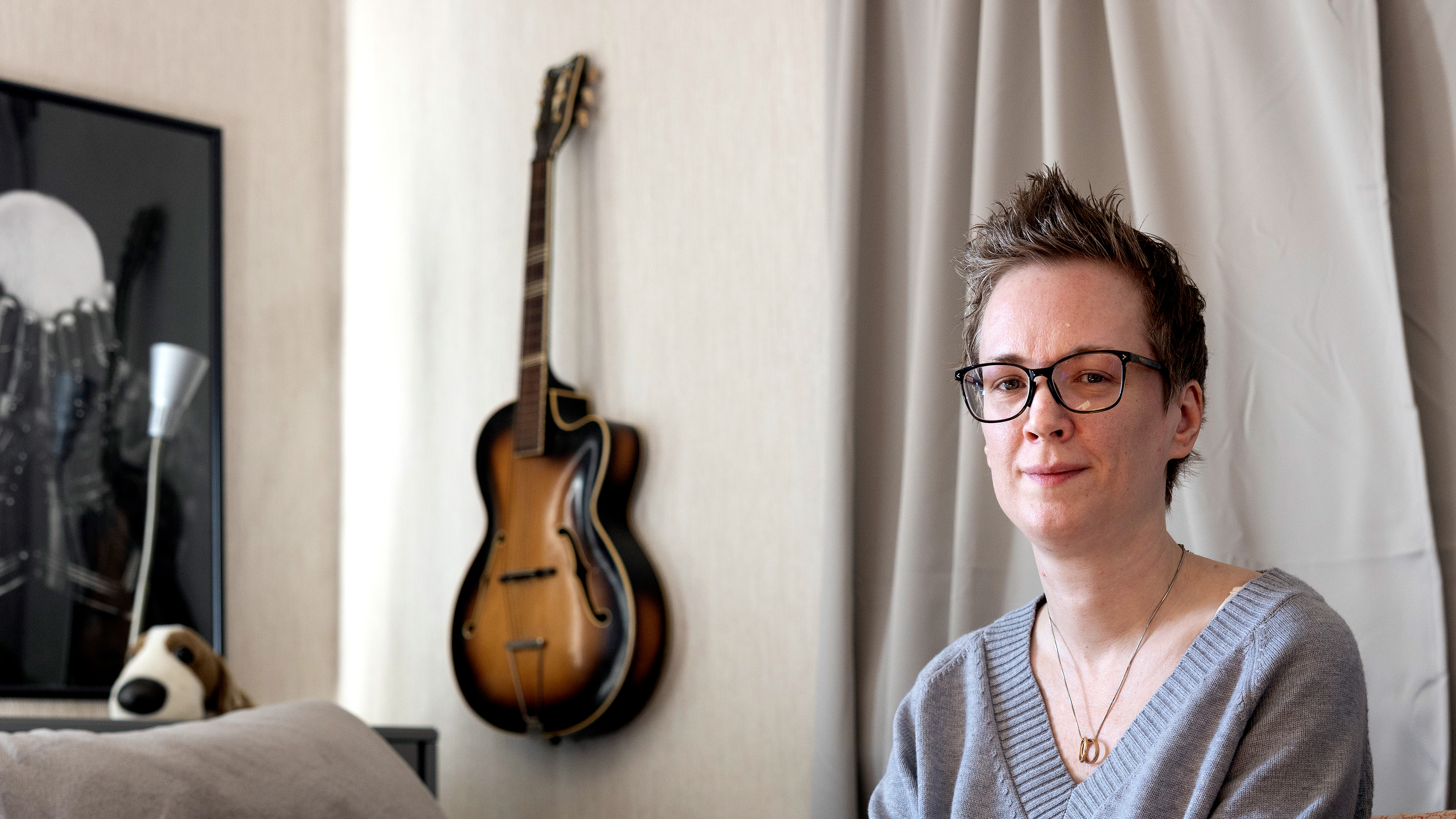 En person med kort hår och glasögon sitter inomhus nära en vägg med en hängande gitarr och ett inramat foto; ett gosedjur ligger på soffan.