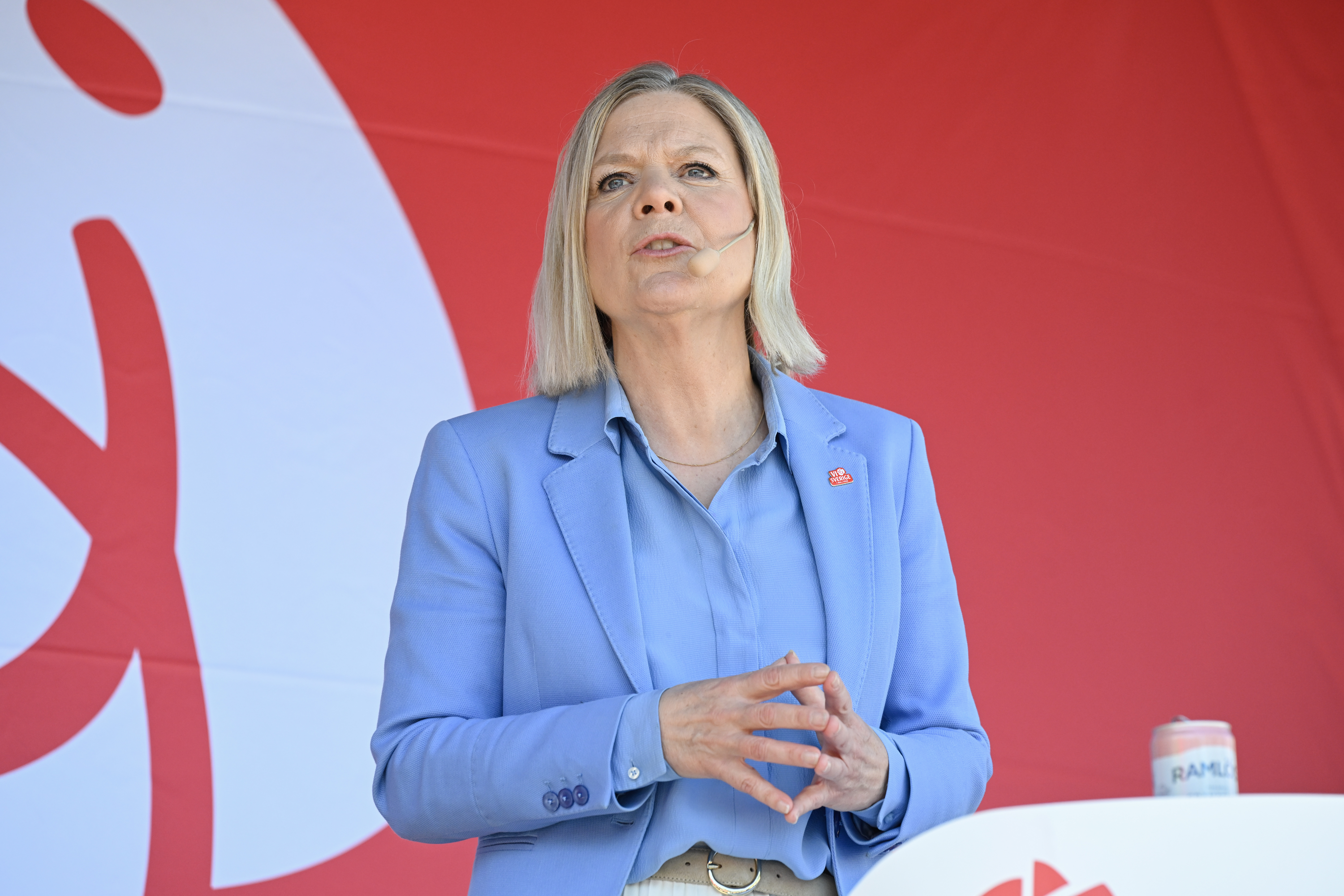 Magdalena Andersson, klädd i ljusblå kostym talar i ett headsetmikrofon framför en röd bakgrund med vit grafik och ett podium bredvid.