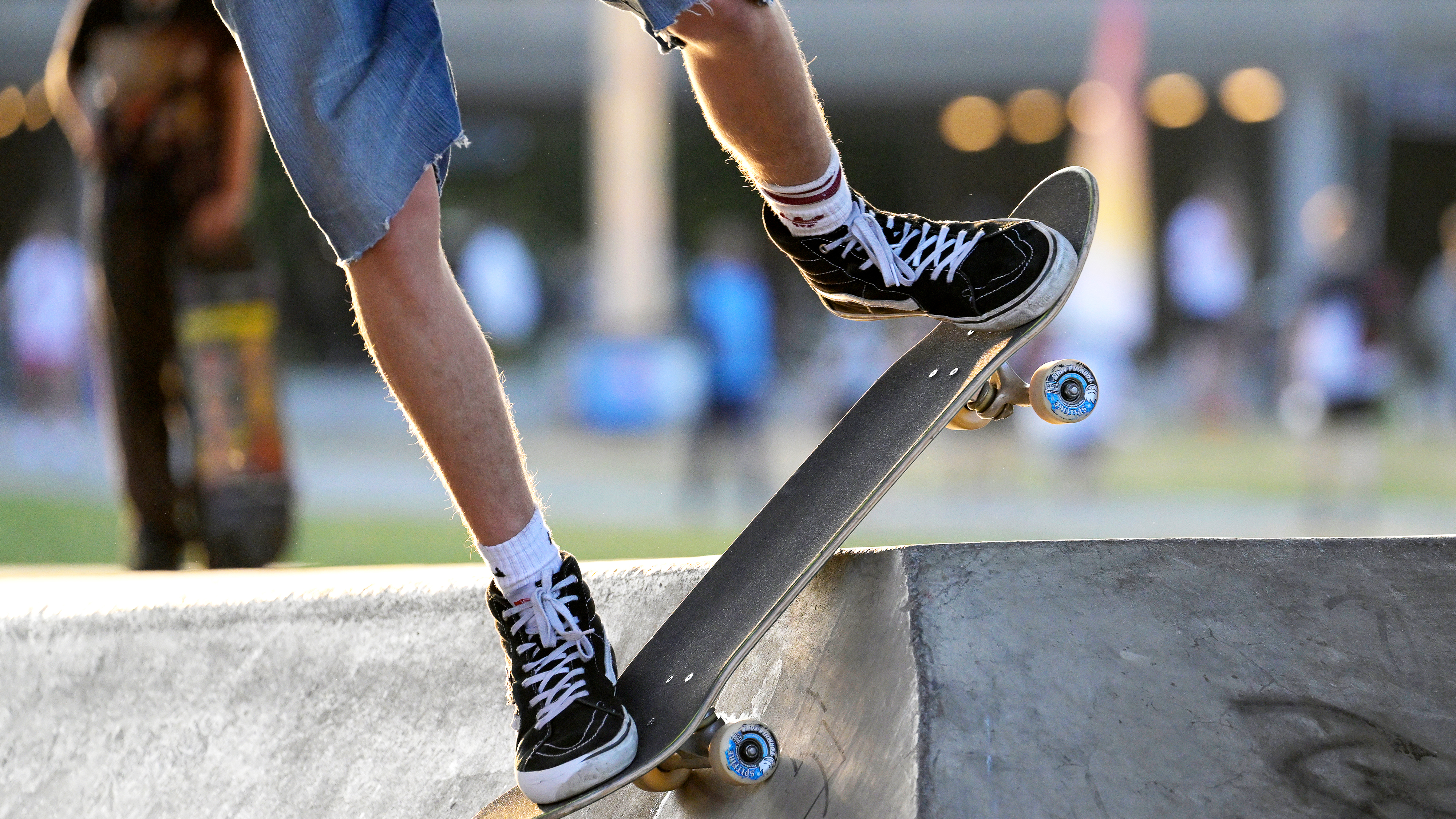 En skateboardåkare med svarta skor och jeansshorts gör ett trick på en betongramp i en skatepark.