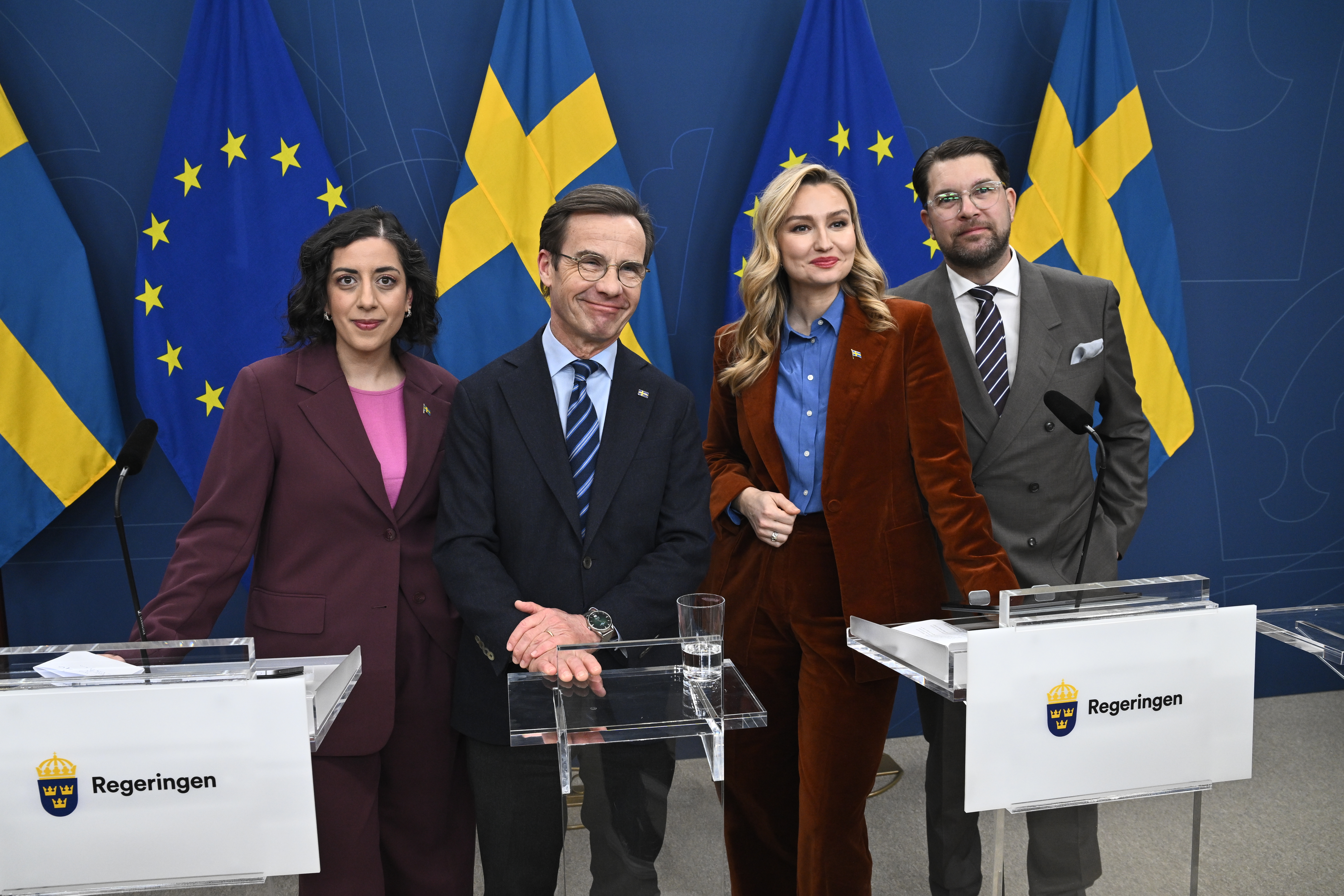 Fyra personer står vid podier med svenska och EU-flaggor i bakgrunden under en presskonferens från regeringen.