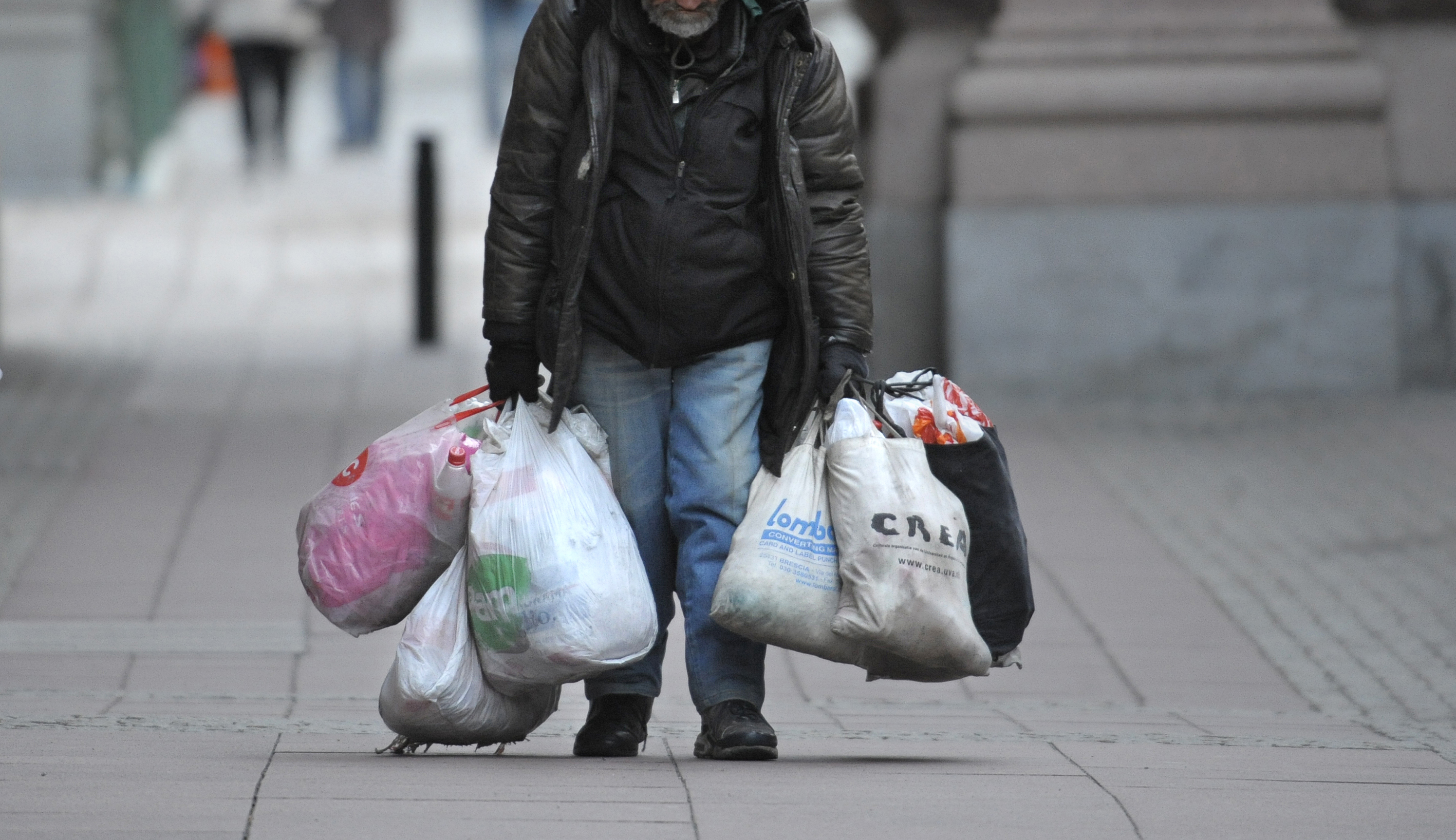 En person går på en trottoar i staden och bär flera stora plast- och tygpåsar, klädd i jacka med huva, jeans och handskar.